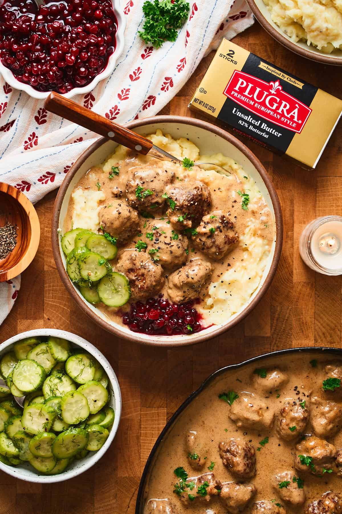 Bowl of Swedish meatballs with creamy gravy over mashed potatoes with lingonberries, cucumbers, candles, linens, and Plugra butter.