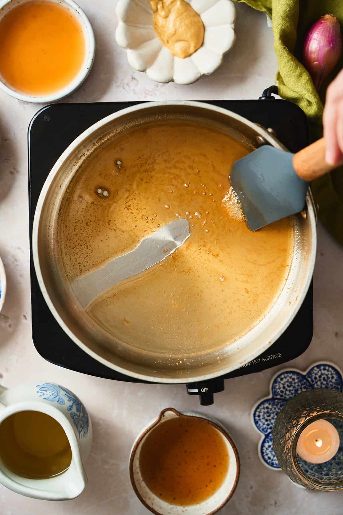 Dragging a rubbers spatula on the bottom of a saucepan surrounded by candles, shallots, and more.