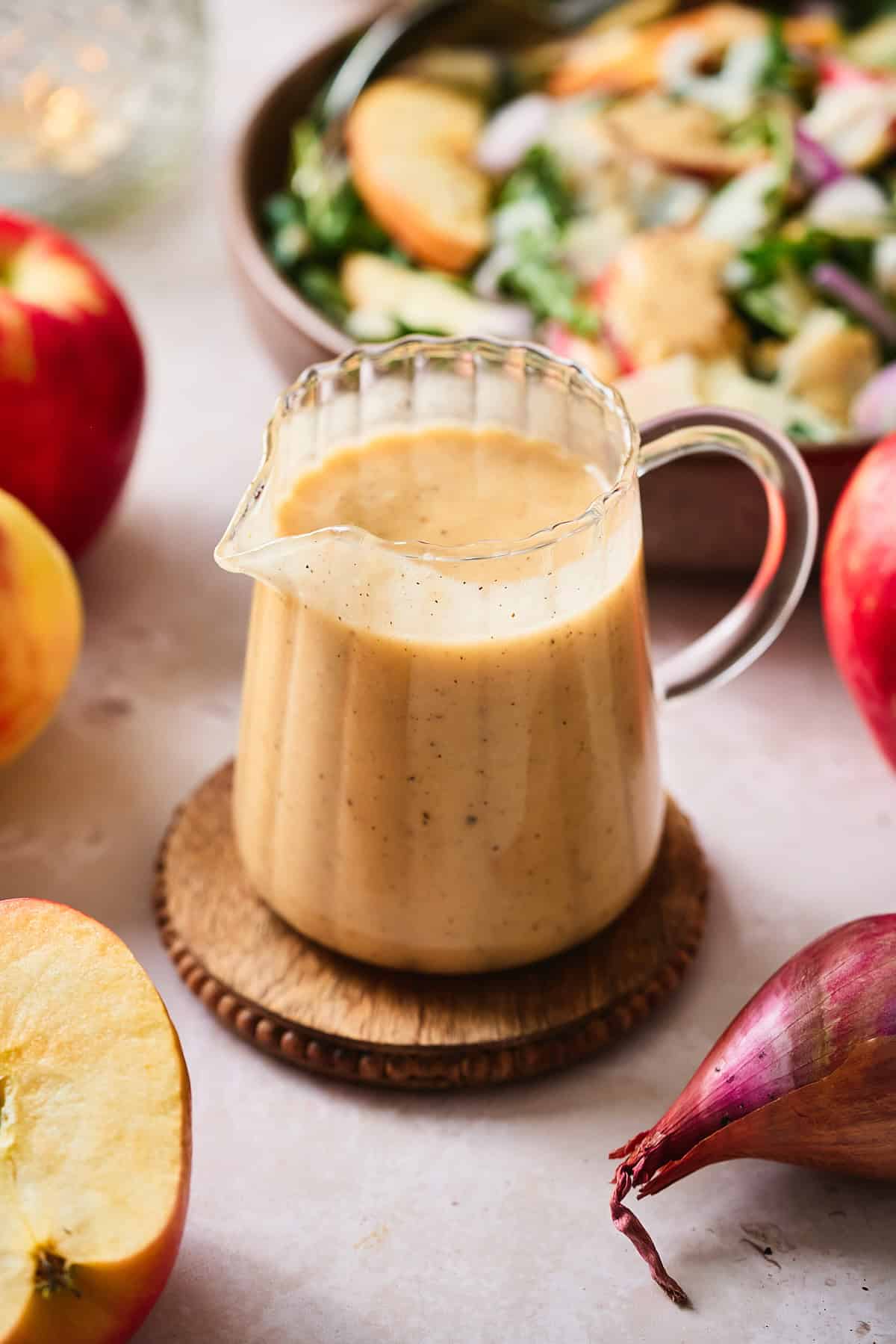 Apple cider dressing in a glass pouring container on a wooden coaster with apples, shallots, and a fresh apple arugula salad behind it. 
