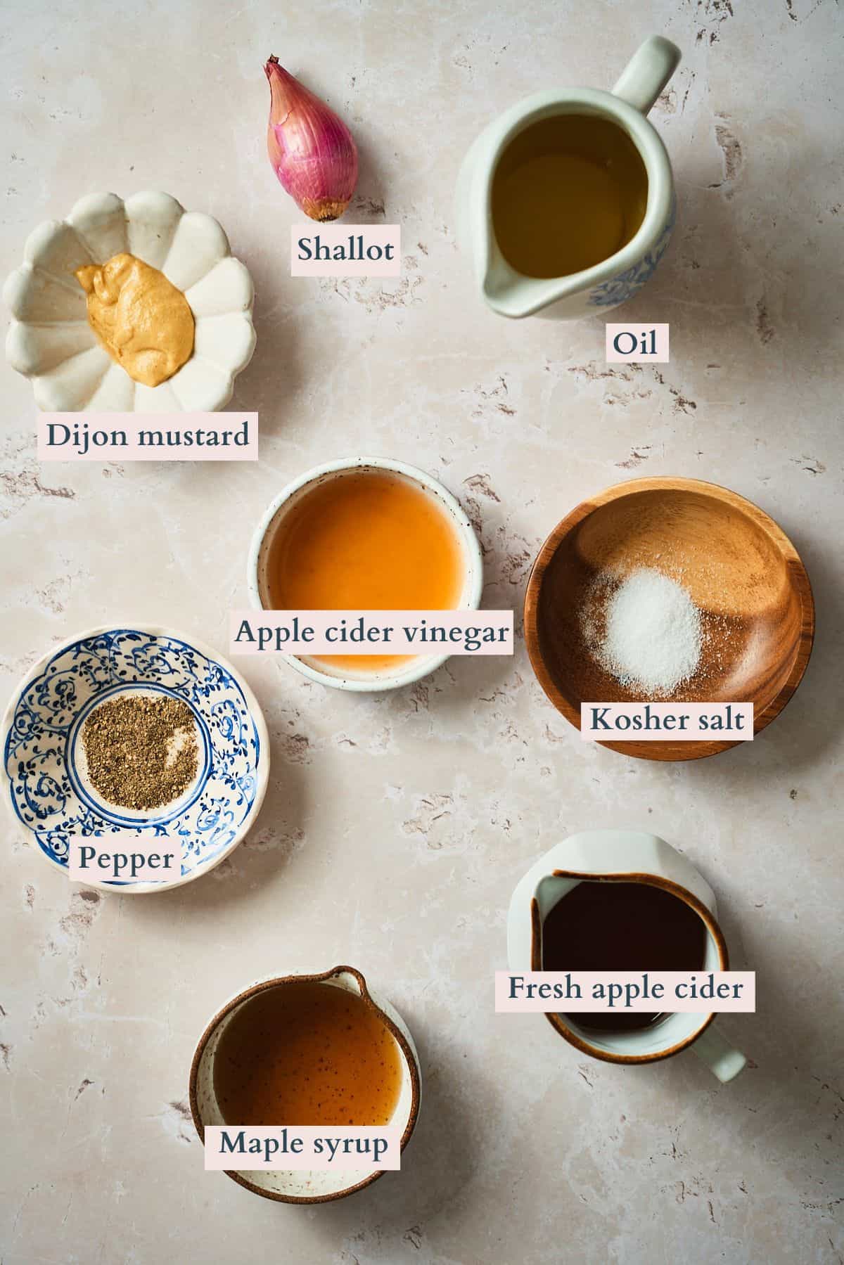 Ingredients to make apple cider salad dressing laid out on a marble counter and labeled to denote each ingredient.