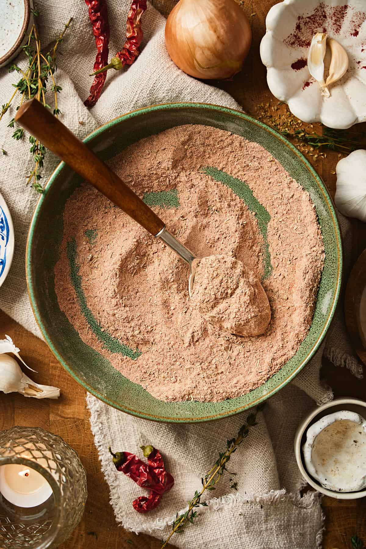 Bowl of seasoning stirred surrounded by garlic, onions, thyme, and a candle.