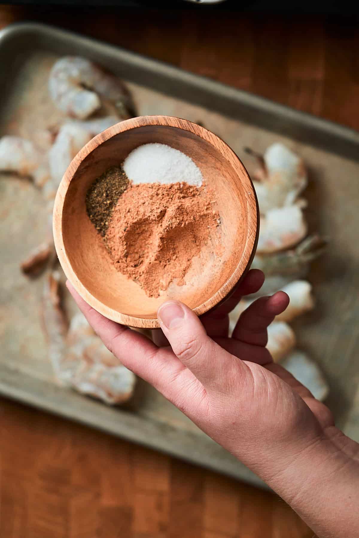 Hand holding salt, pepper, and Cajun seasoning in a bowl over raw shrimp.