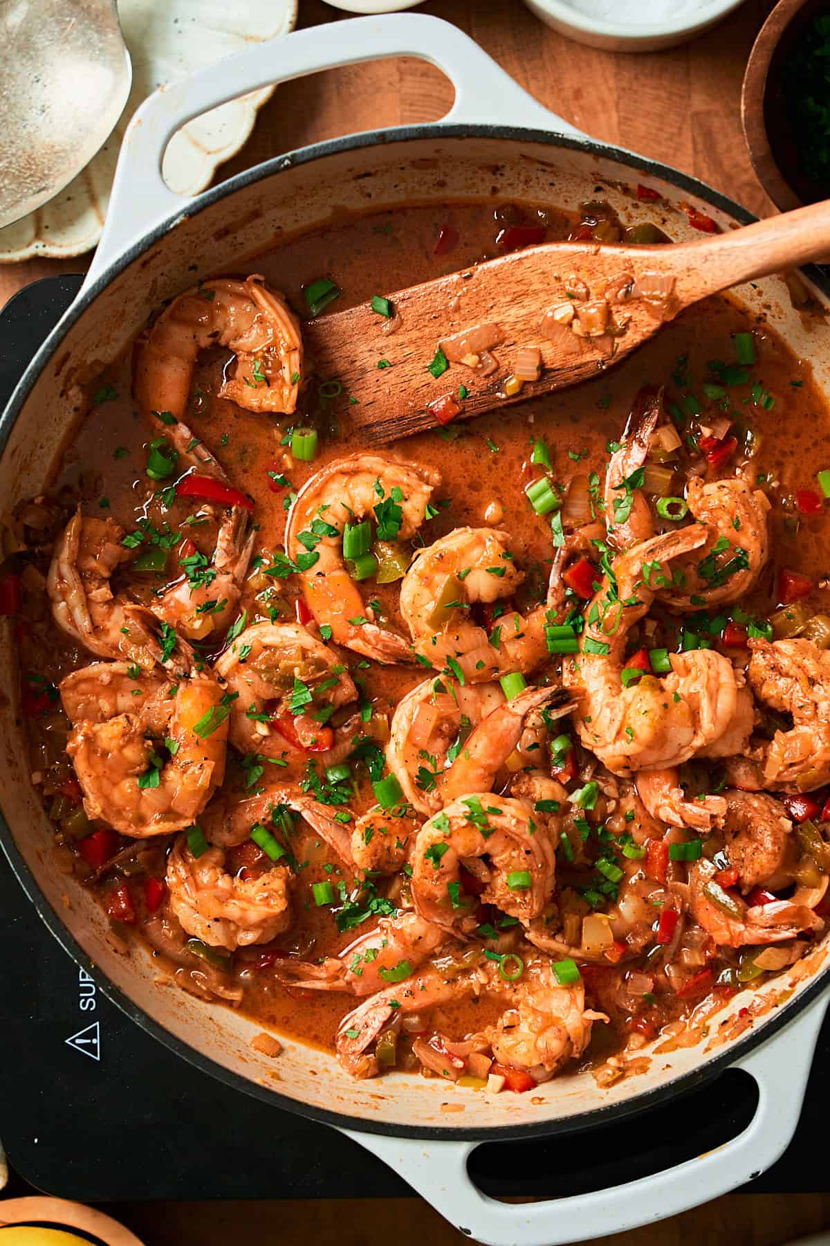 Close up shot of glossy coated shrimp with green onions and parsley in a skillet with a wooden spoon.