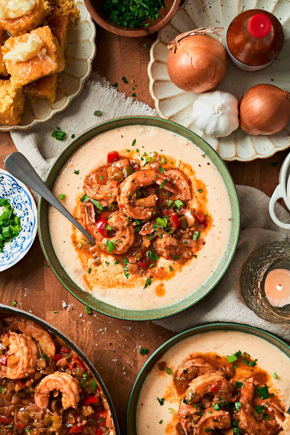 Ovehead shot of a bowl of creamy shrimp and grits surrounded by a skillet, cornbread, onions, parsley, and a candle.