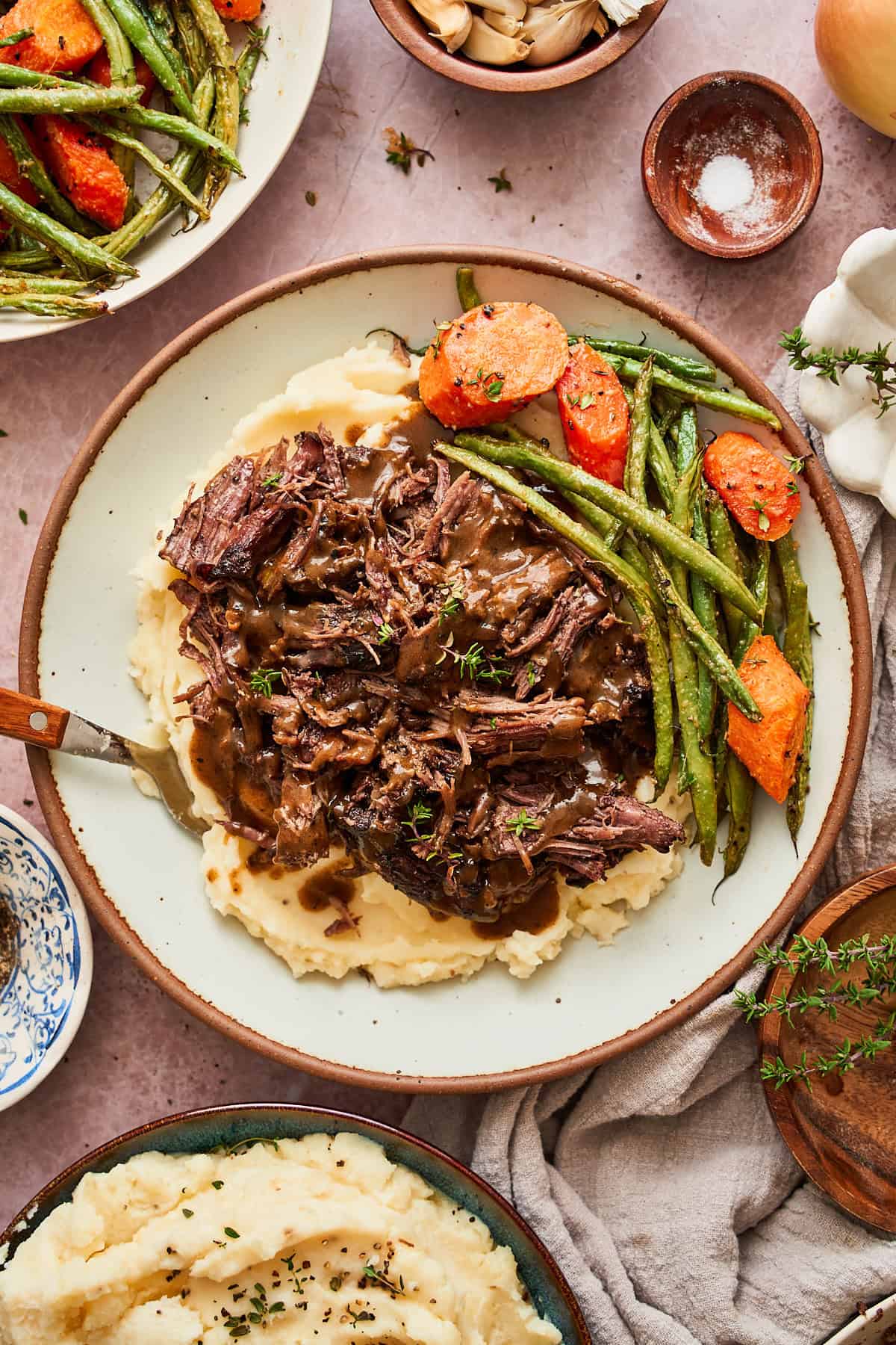 Plate of mashed potatoes topped with shredded beef, with gravy, green beans, carrots and surrounded by onion, garlic, and herbs.