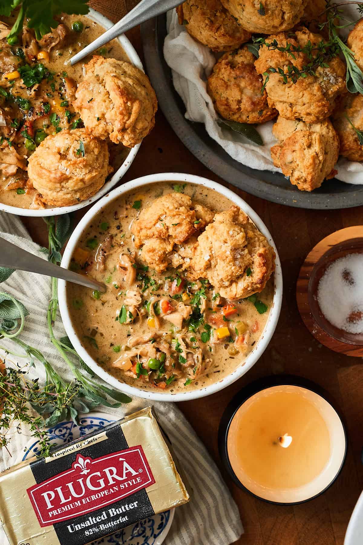 Flat lay shot of two bowls of creamy chicken pot pie soup topped with herbed drop biscuits with plugra butter nearby, fresh herbs, salt, candles, and drop biscuits.