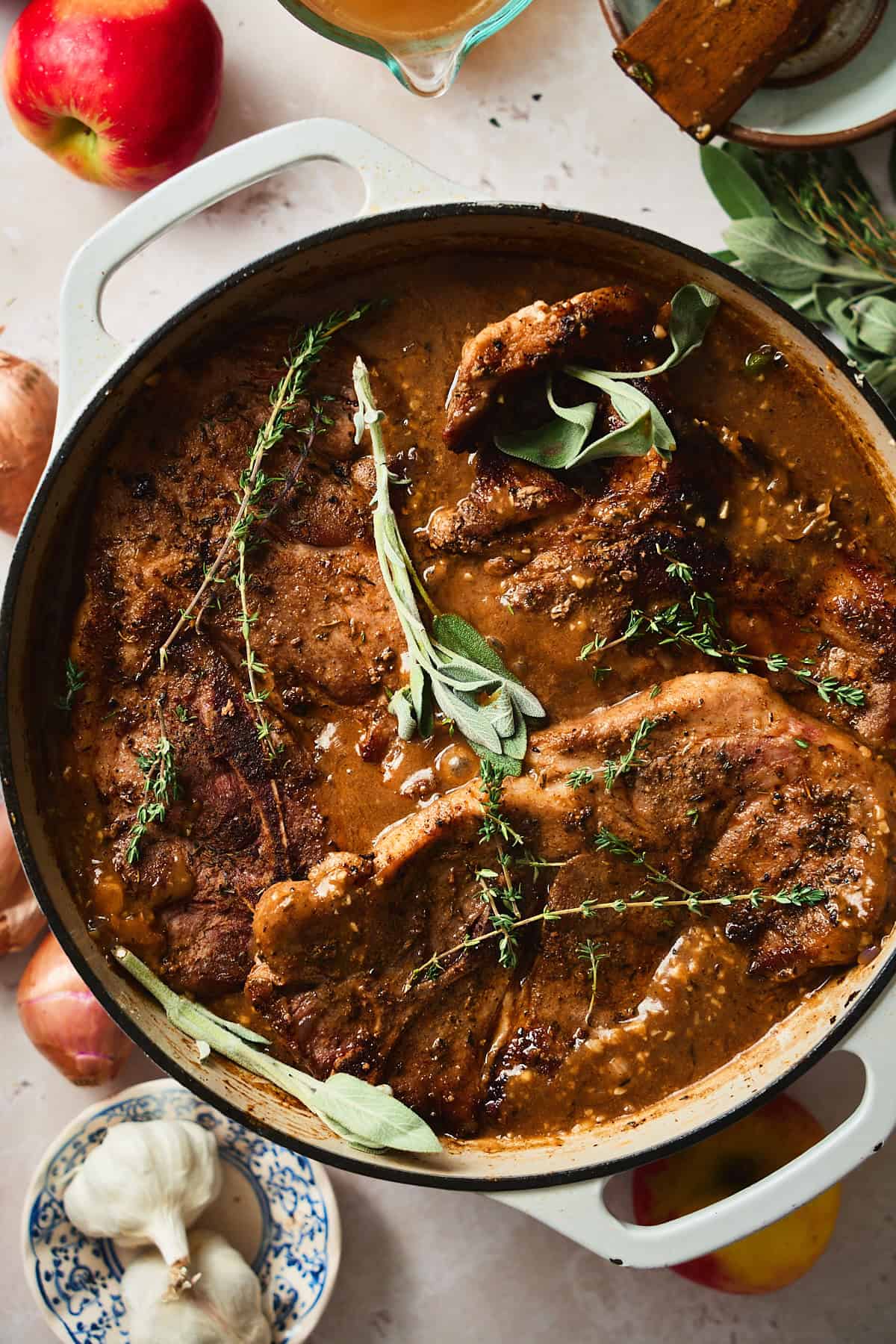 Pork steaks nestled into a pan with gravy in it, with fresh thyme and sage added.