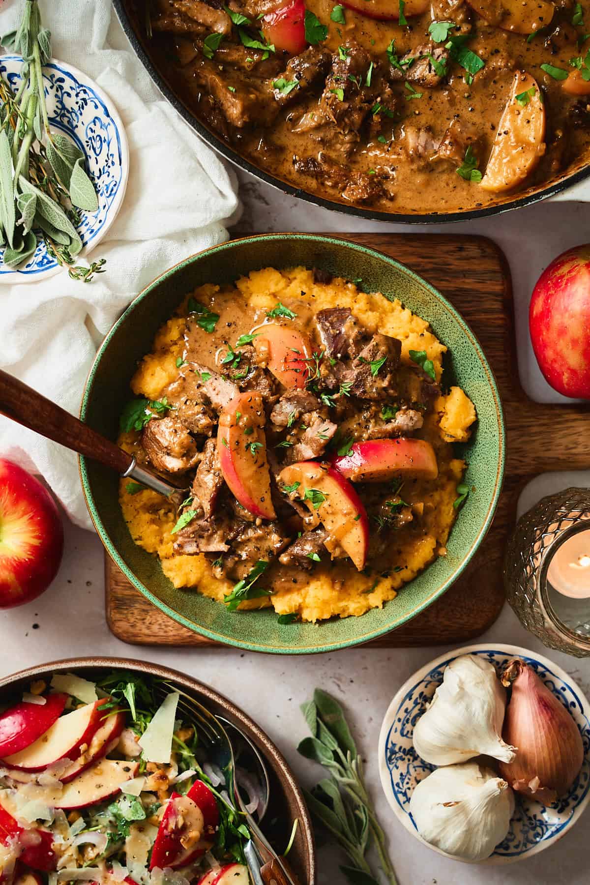 Stunning photo of creamy pork and apple dish over a bed of mashed rutabaga with an apple arugula salad, fresh herbs, and apples.