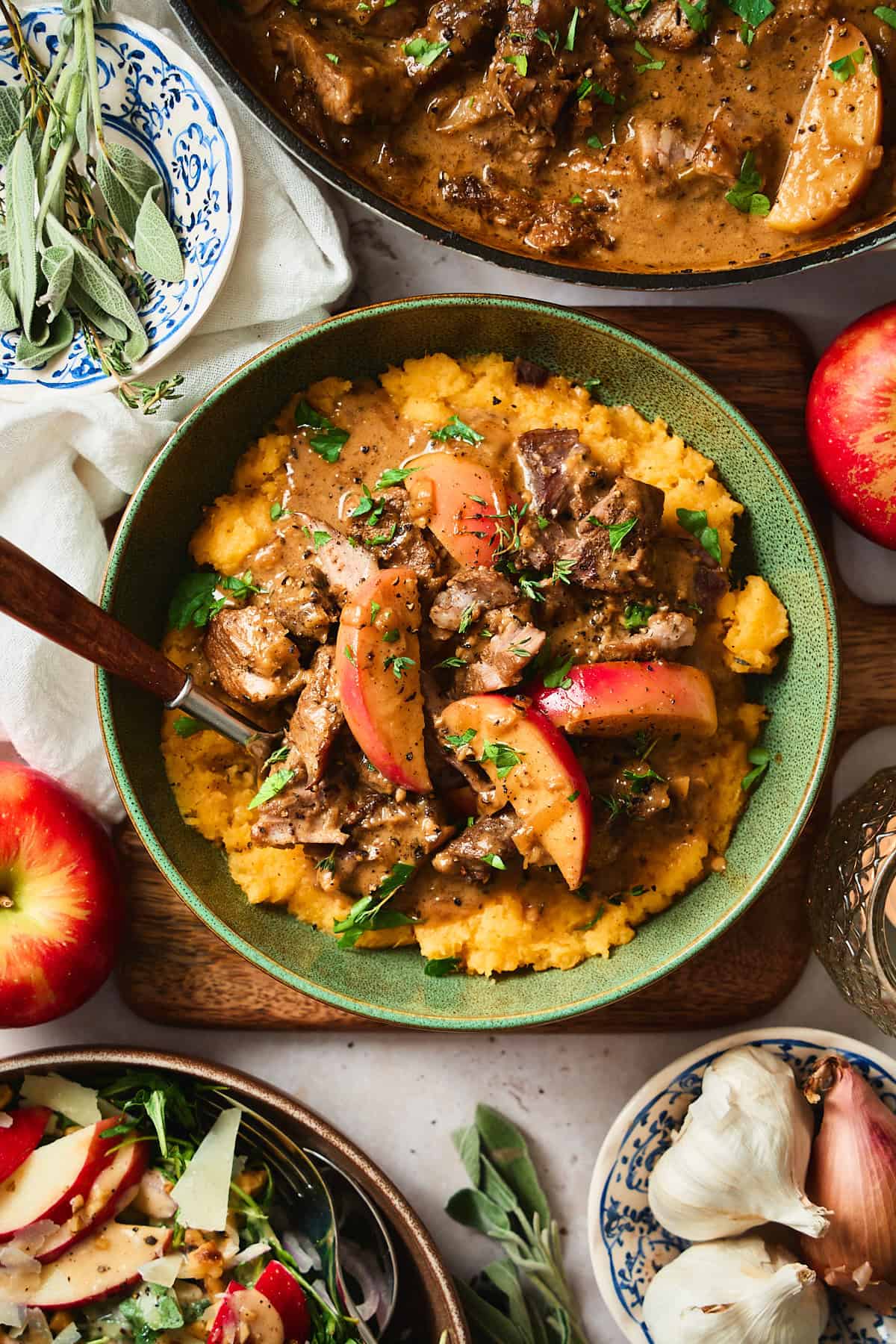 Gorgeous colorful shot of a plate with mashed rutabaga, creamy braised pork with apples, with a salad, fresh herbs and garlic nearby. 