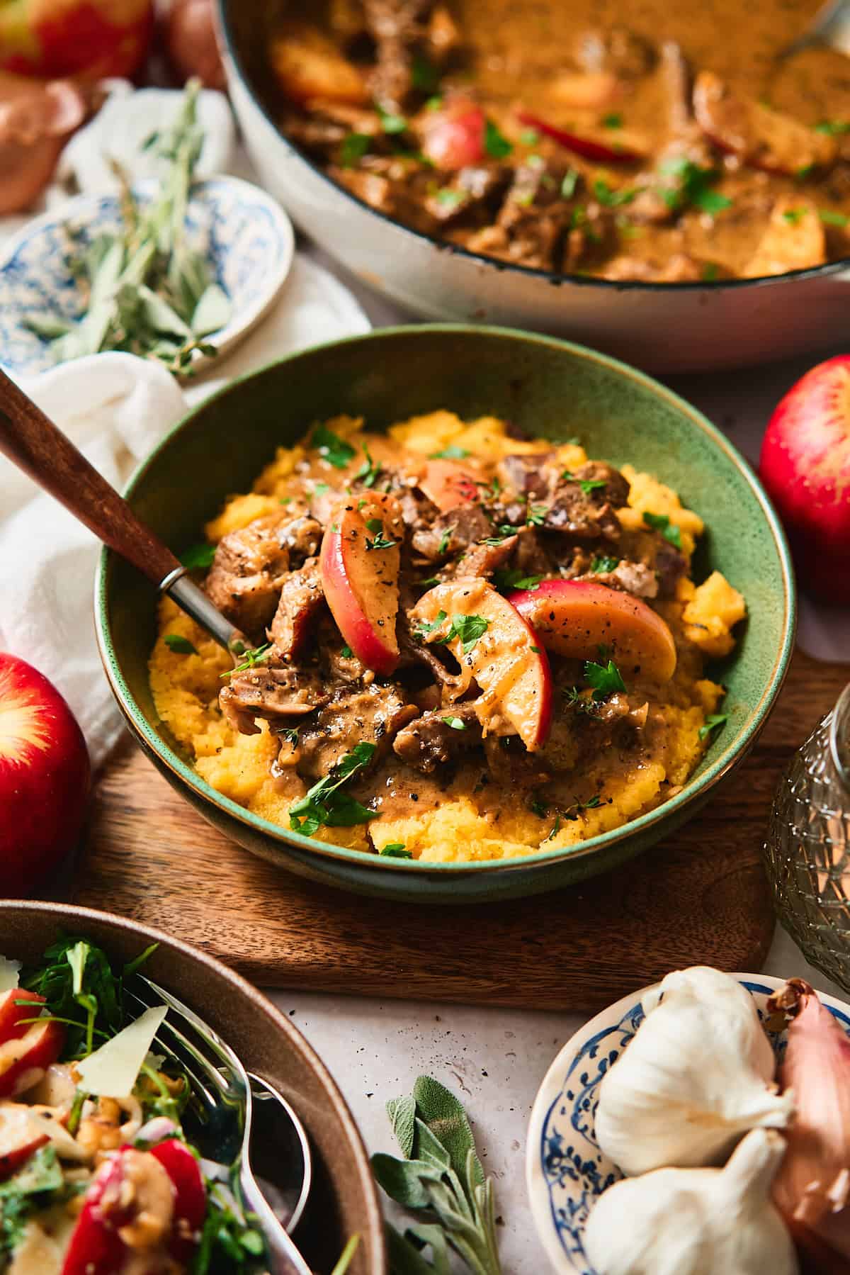 Beautiful 45-degree shot of a green bowl filled with mashed rutabaga and a creamy smothered pork and apples surrounded by a salad, garlic, shallots, fresh apples, a candle, and herbs.