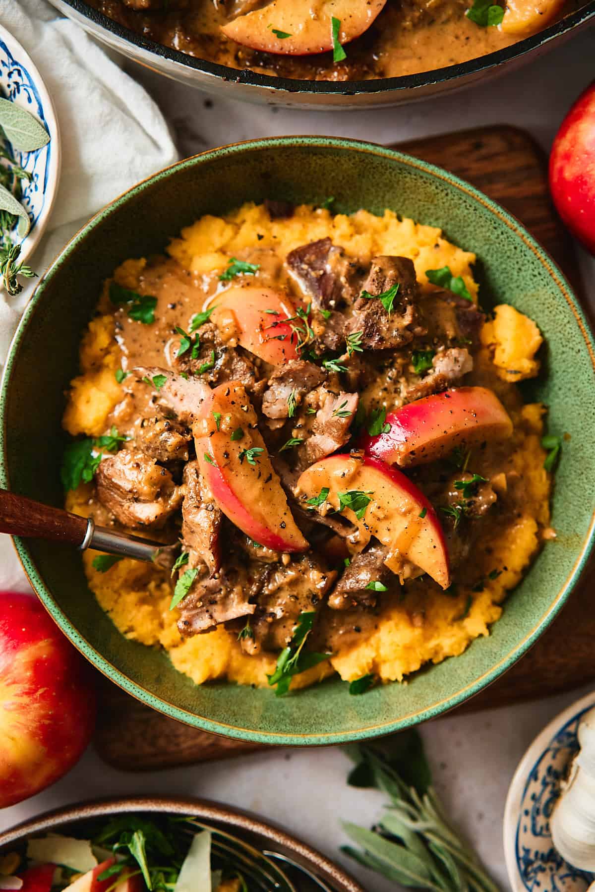 Close up shot of a green bowl filled with orange mashed rutabaga, creamy braised pork and apples with a creamy gravy and a salad nearby.
