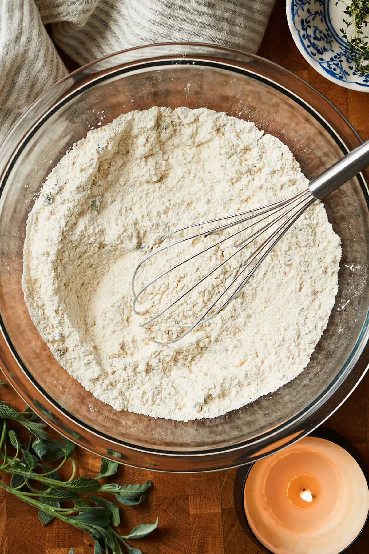 Whisked ingredients to make herbed drop biscuits in a bowl with a whisk, fresh herbs, and a candle nearby.