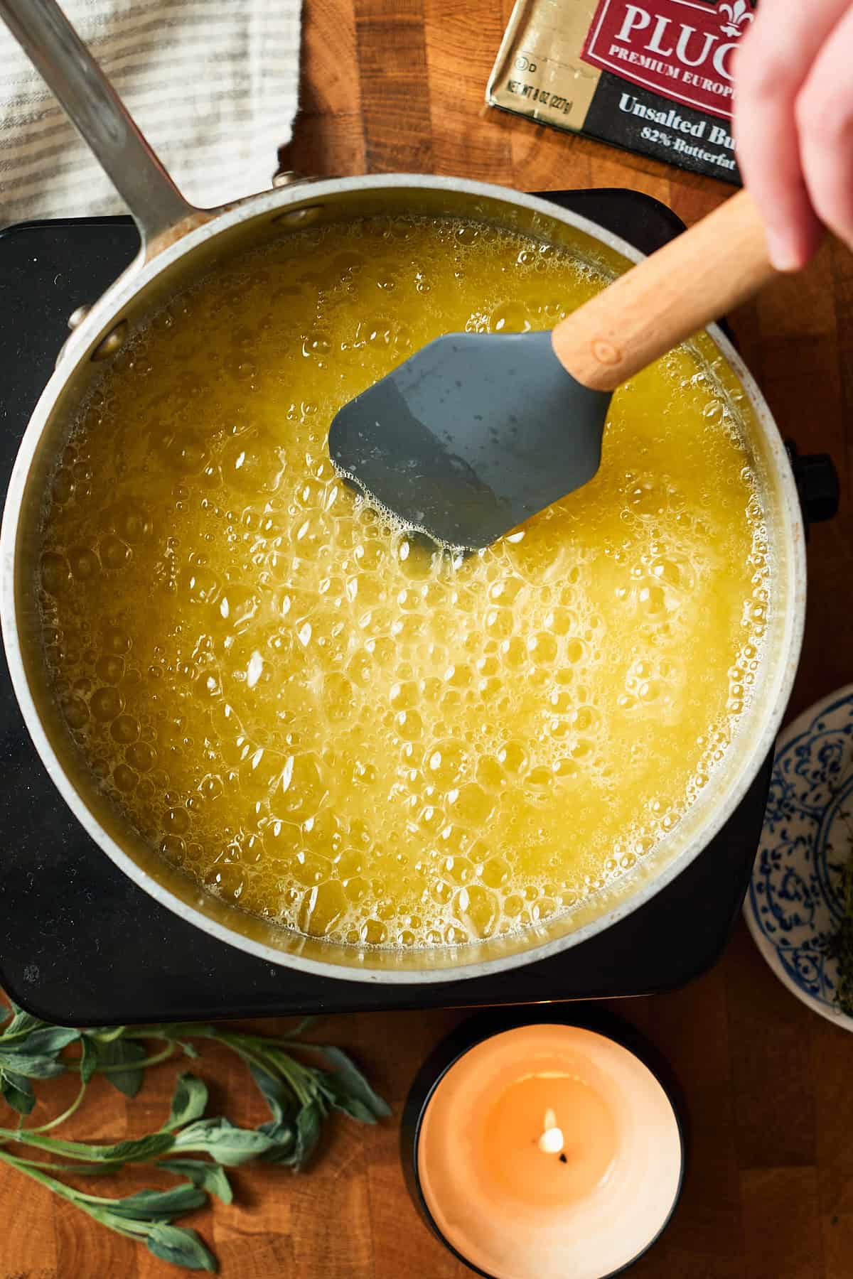 Butter melting in a metal saucepan with a rubber spatula in the saucepan surrounded by fresh herbs and a candle.