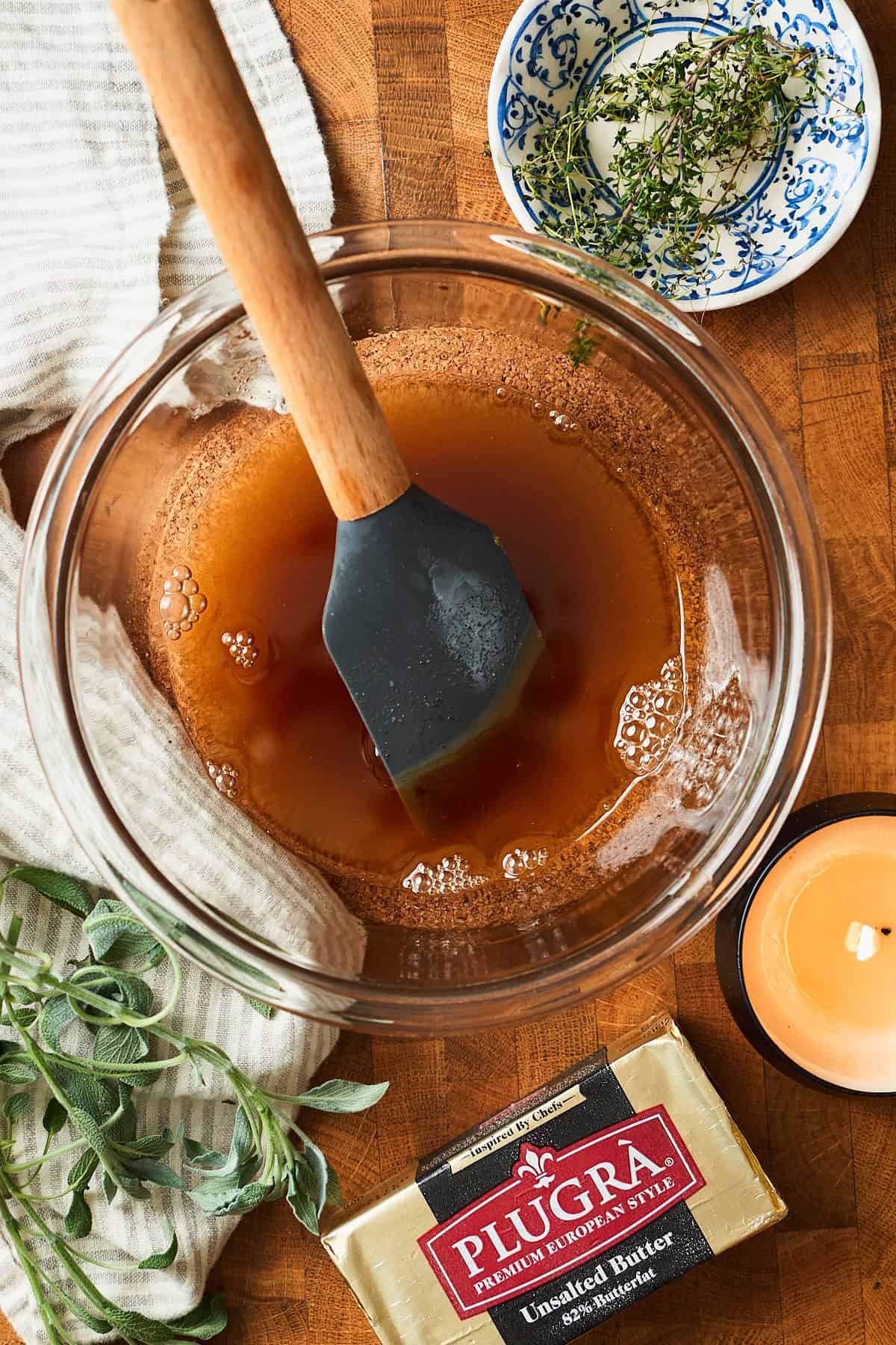 Brown butter in a bowl cooling off surrounded by fresh herbs and a candle.