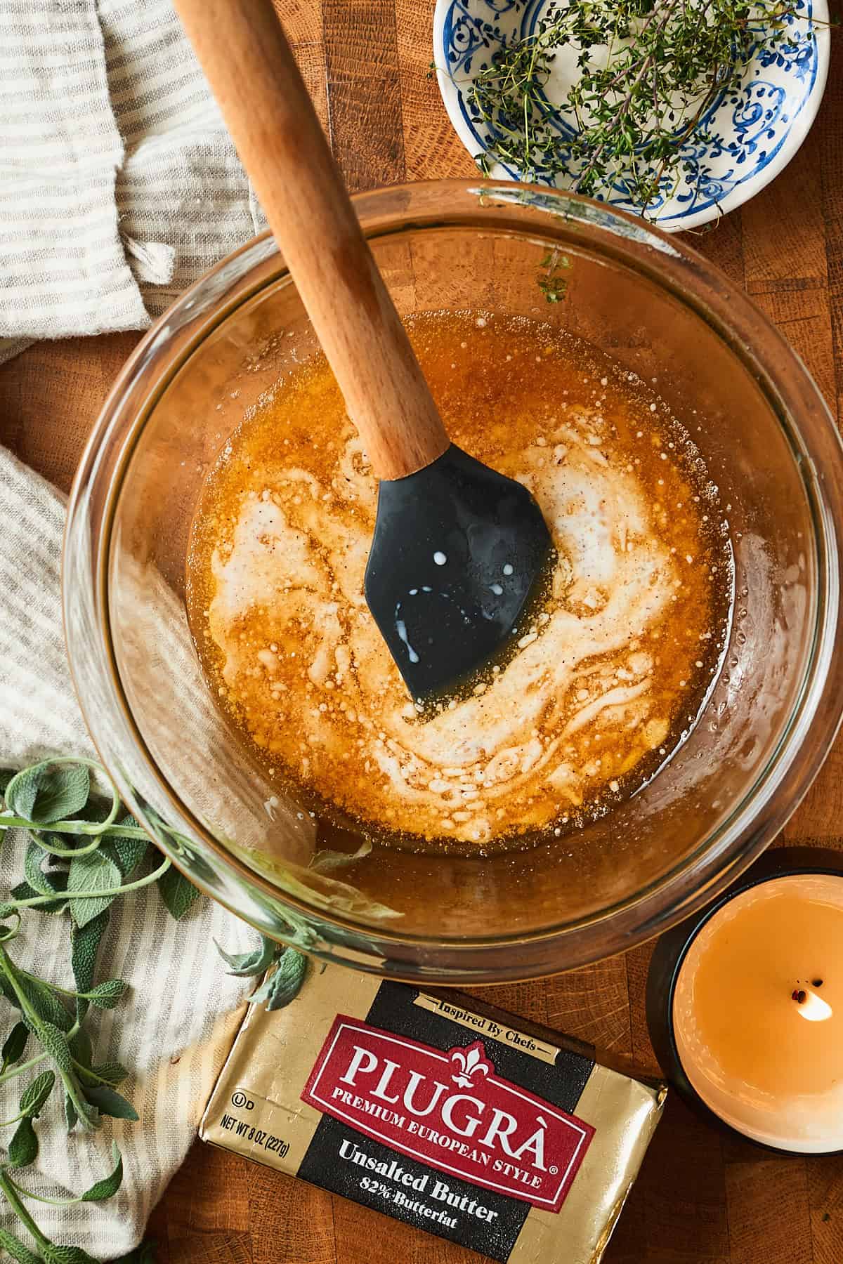 Buttermilk and brown butter being combined in a bowl with a candle, a stick of plugra, and herbs nearby.