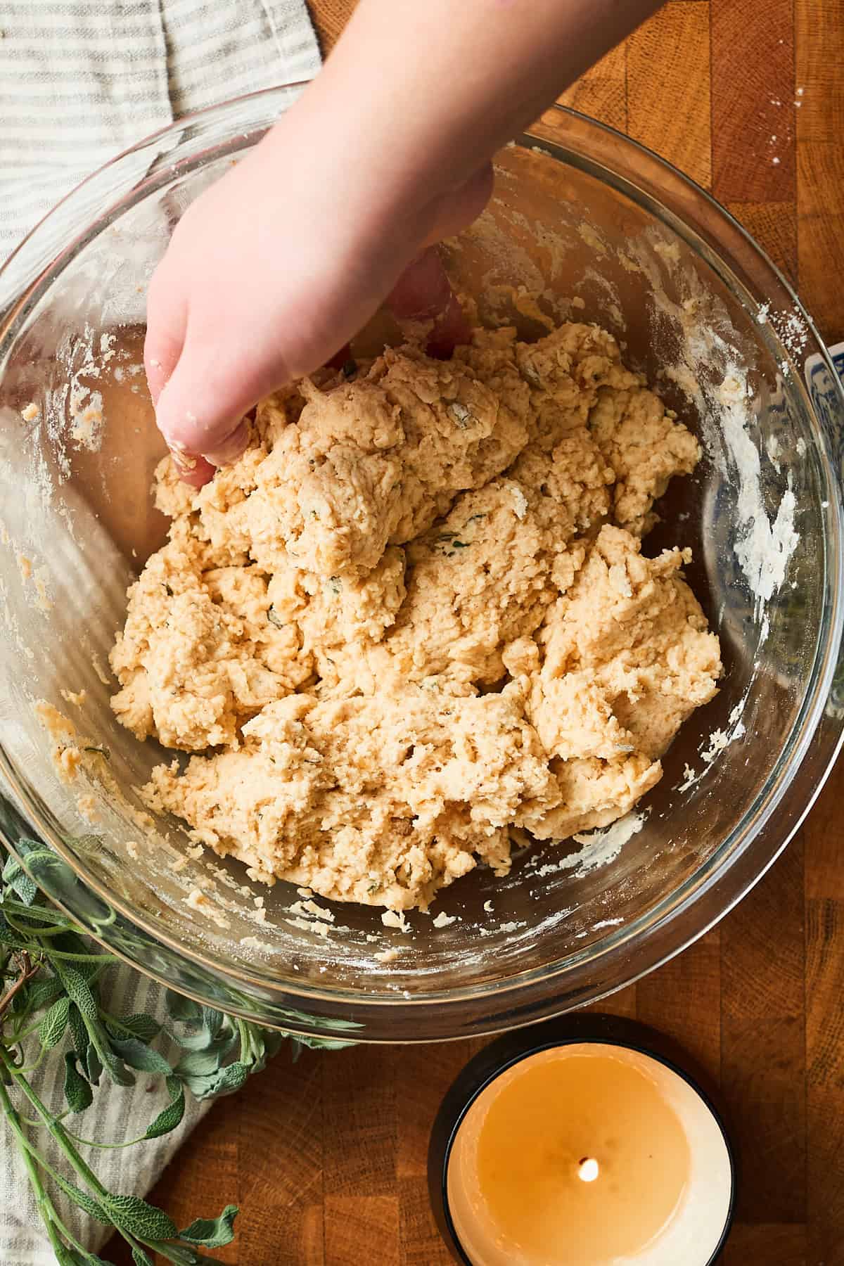 Person using their hands to mix together wet and dry ingredients for drop biscuits in a glass bowl surrounded by herbs and a candle.