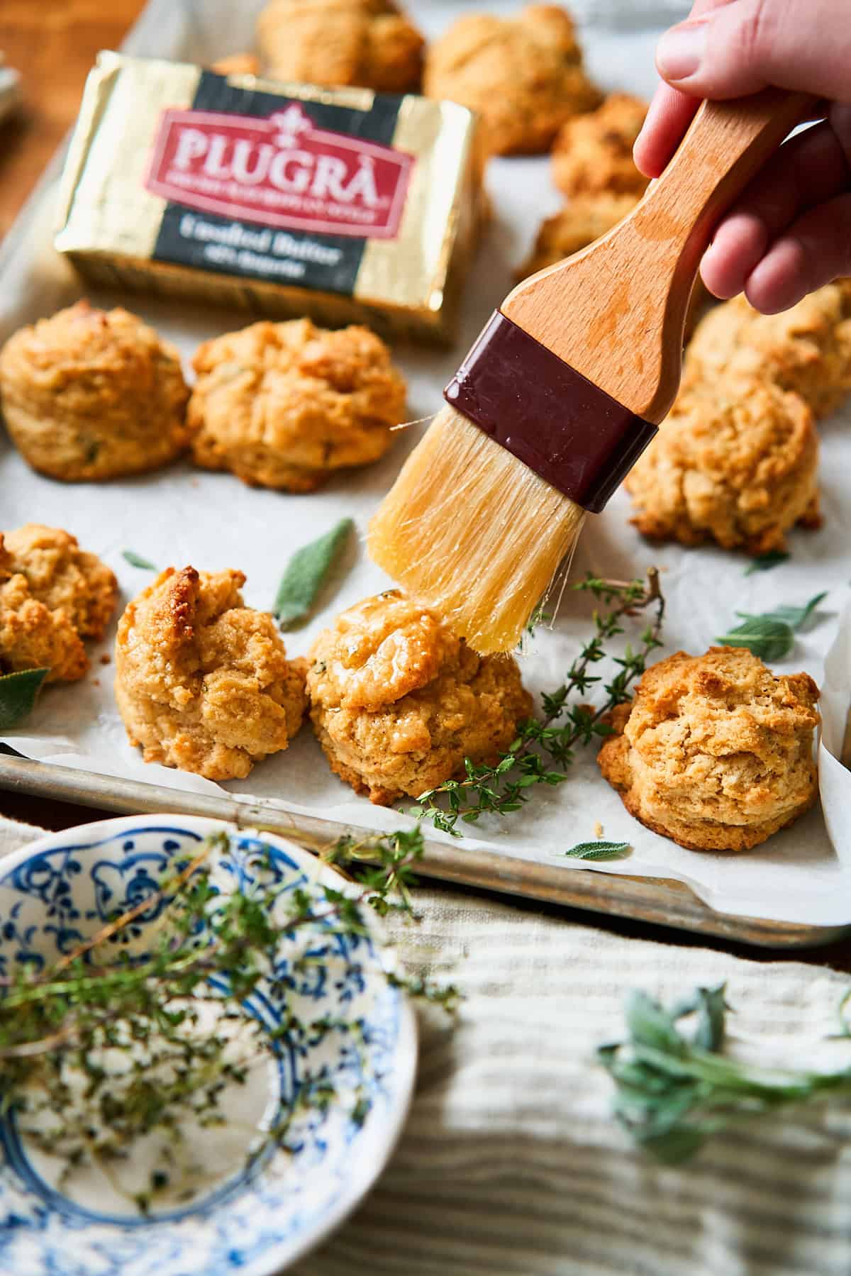 Brushing brown butter drop biscuits with more melted butter, surrounded by fresh herbs and plugra butter nearby.