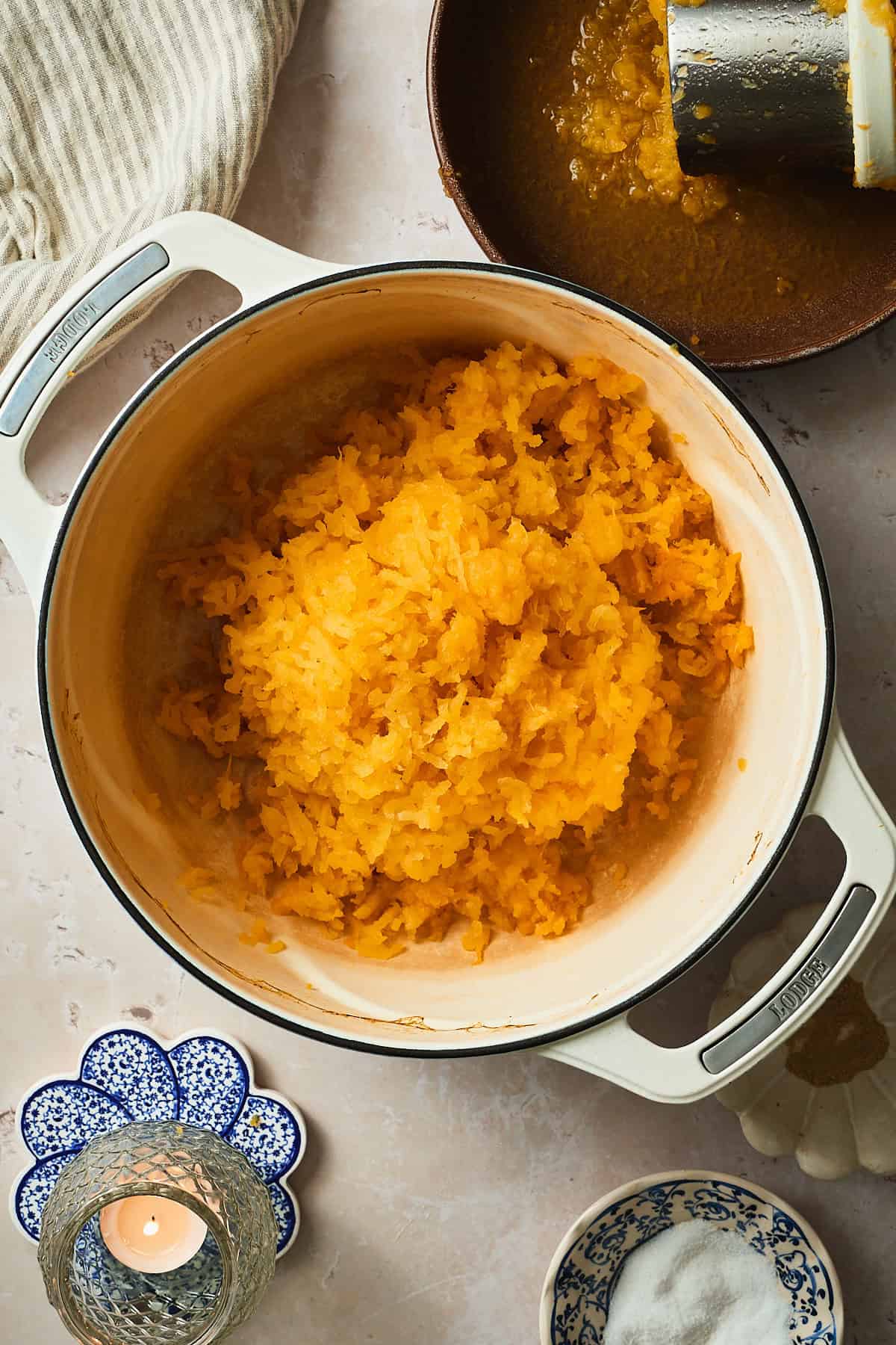Riced rutabaga in a pot with a potato ricer nearby, and a candle on a blue coaster.