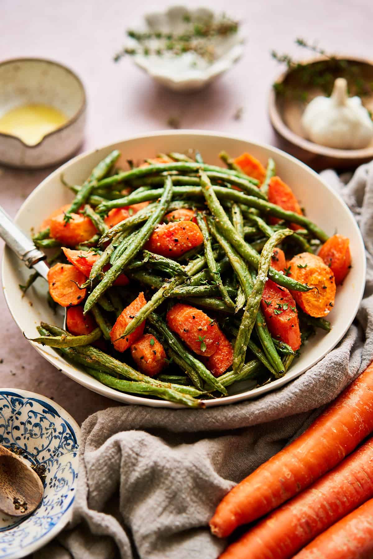 45 degree angle of a bowl of green beans and carrots surrounded by garlic, fresh herbs, and fresh carrots,