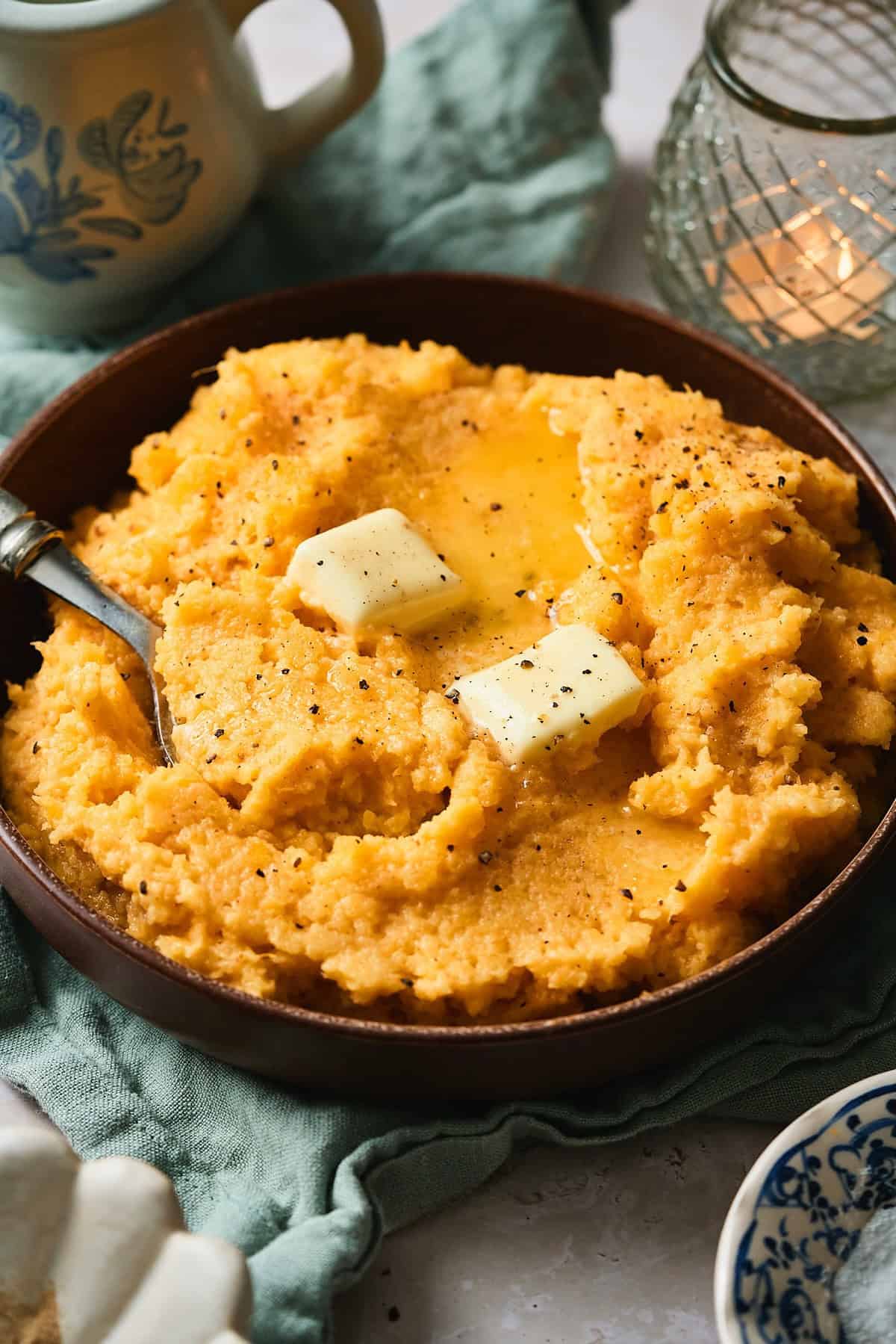 45 degree shot of a bowl with mashed rutabaga topped with melting butter with a twinkling candle in the background.