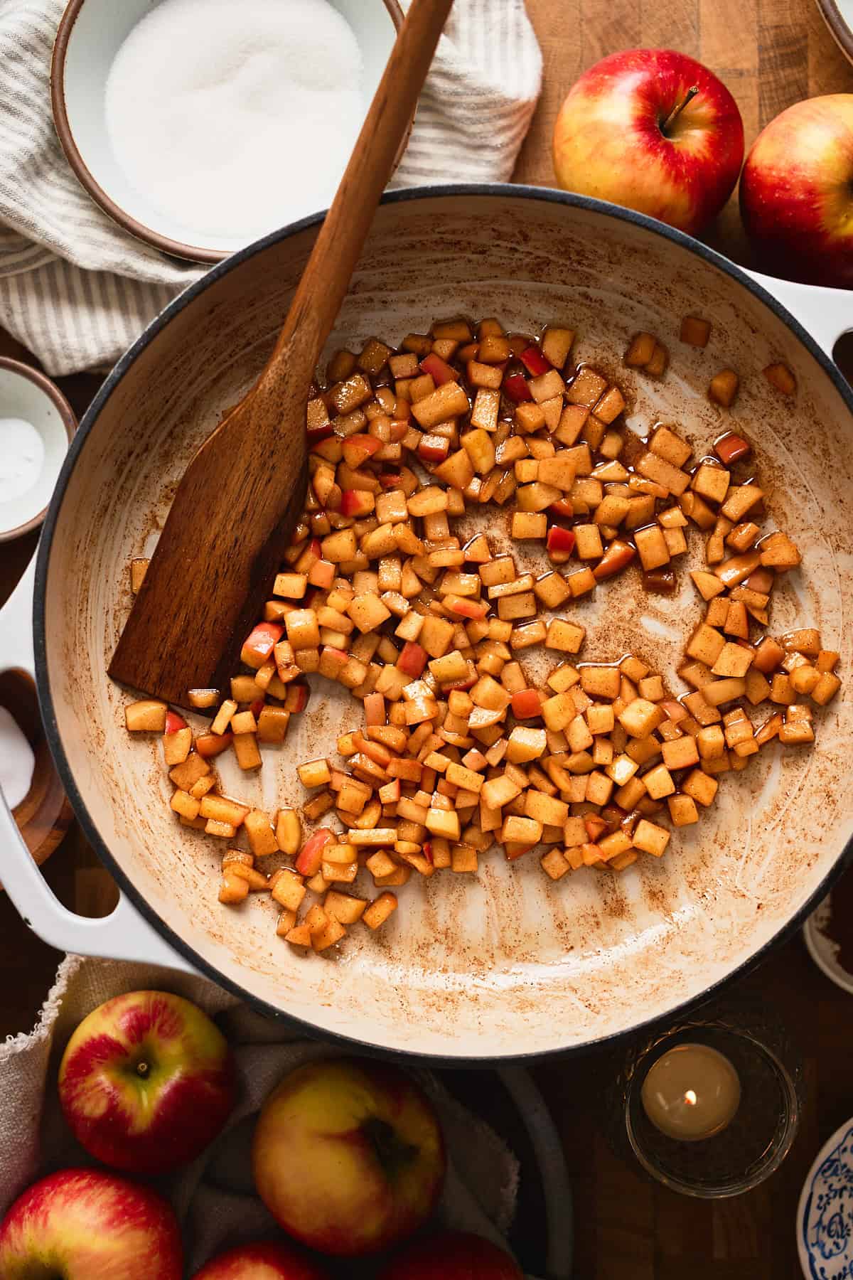Cooked apple cinnamon chunks in a enameled skillet surrounded by fresh apples, candles, and whole spices.