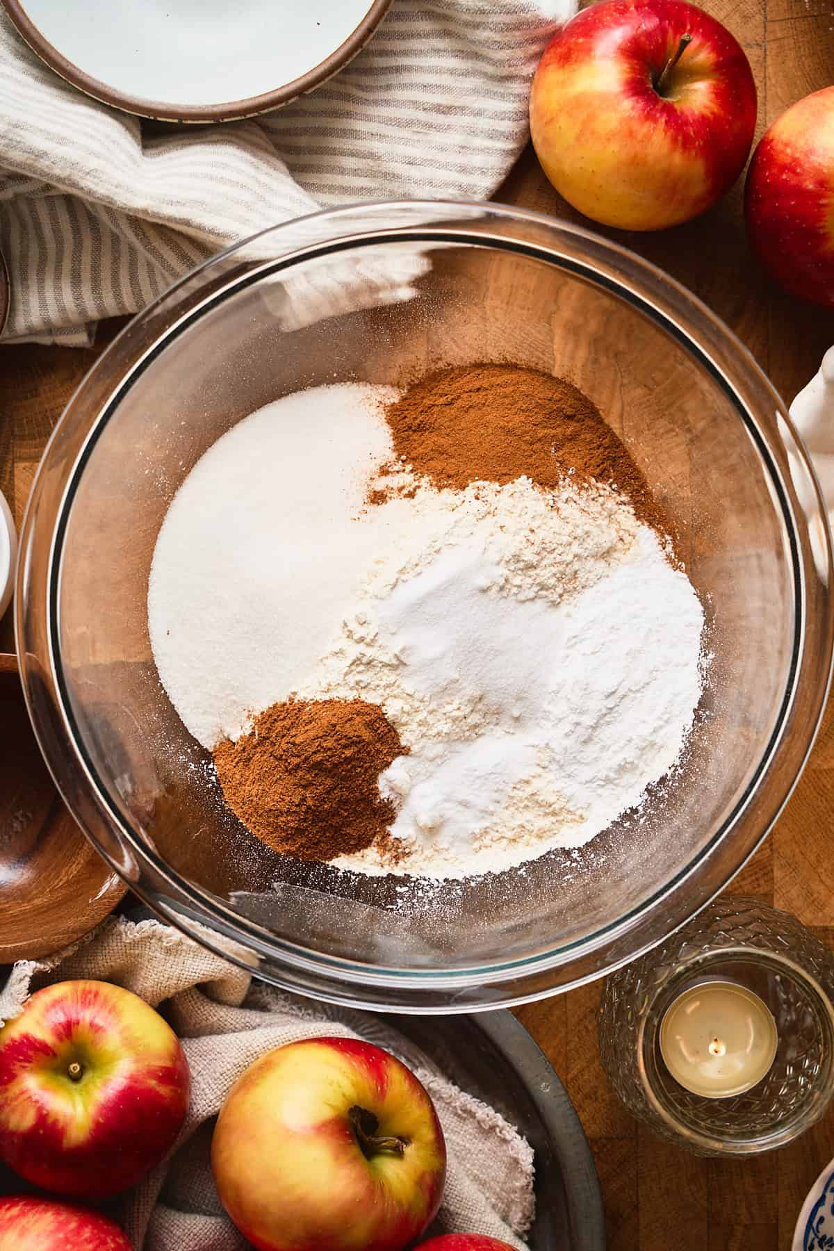 Flour, sugar, spices, salt, and leavening agents in a mixing bowl surrounded by apples, candles, and linens.