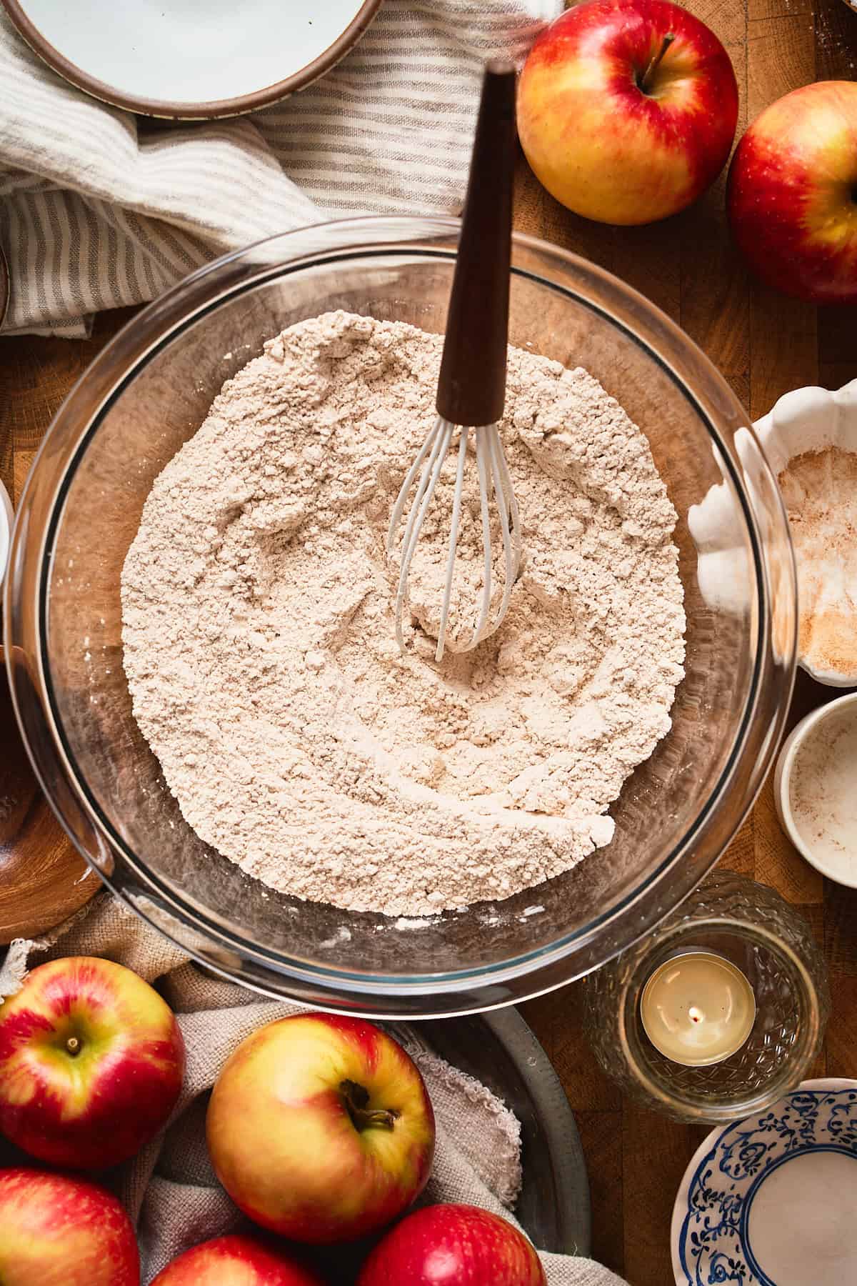 Whisked flour and dry ingredients in a bowl surrounded by apples, candles, and linens for a warm cozy feel.