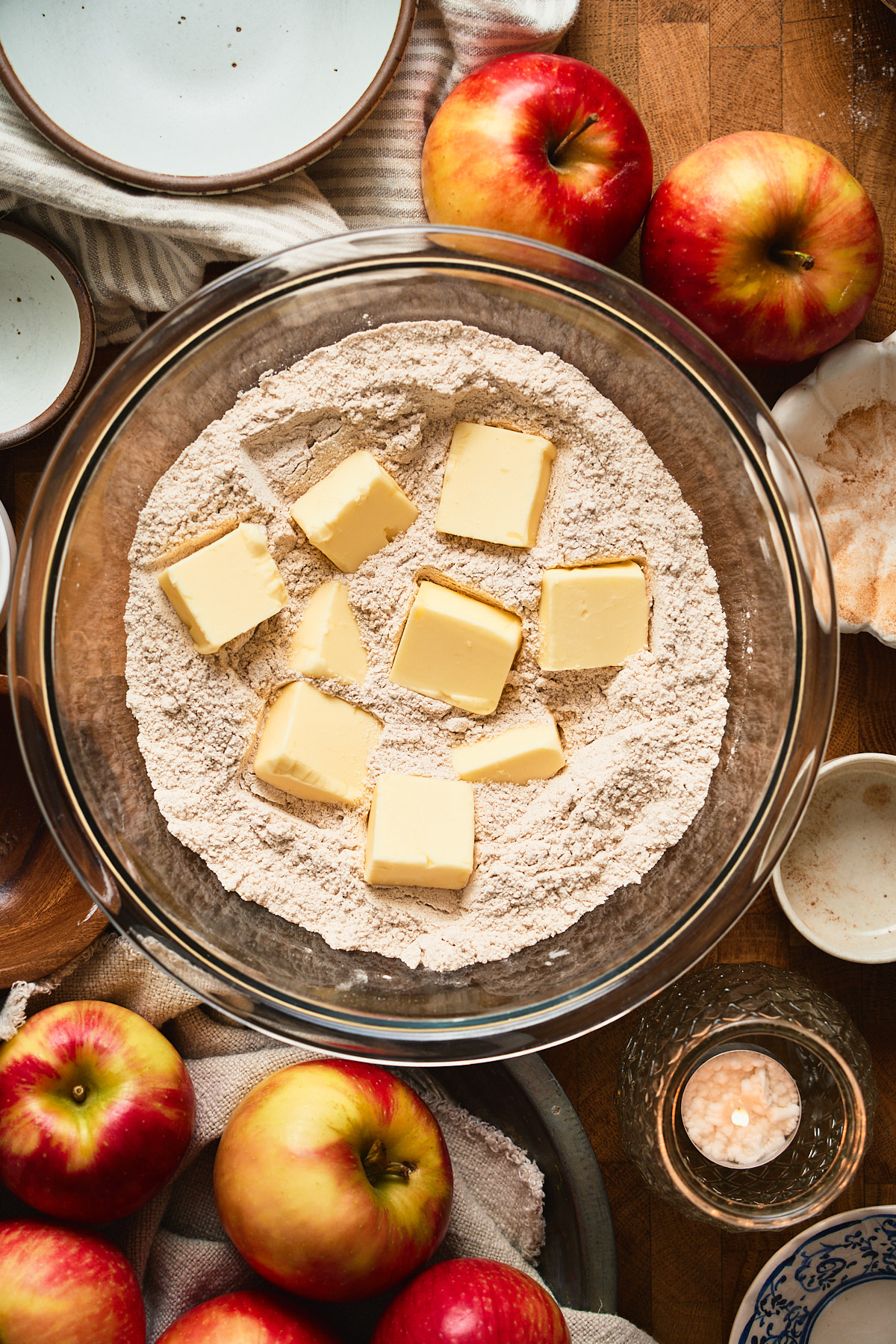 Butter pieces in a bowl of flour surrounded by apples, candles, and small bowls of different sizes.