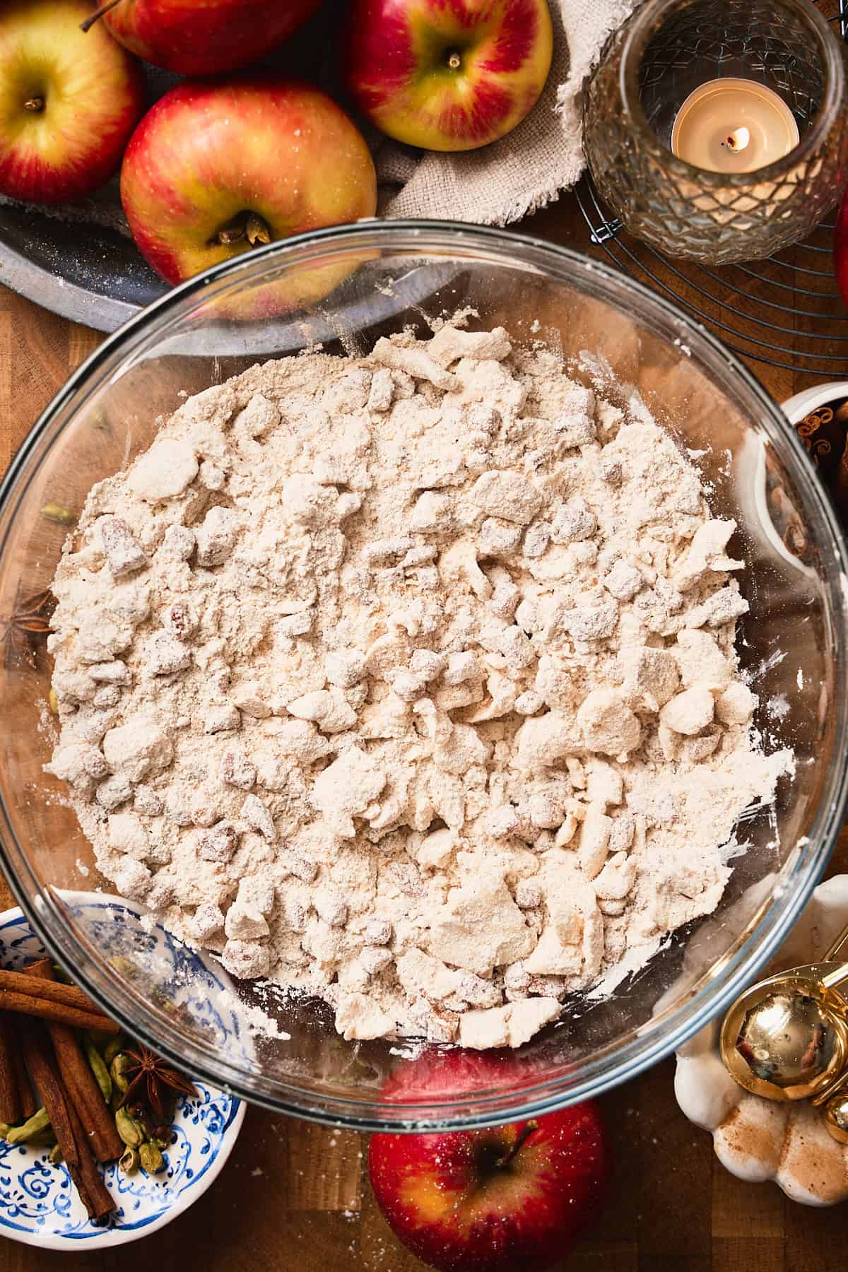 Dough beginning to form with butter pieces and apple chunks covered in flour surrounded by fresh apples, spices, candles, and more.