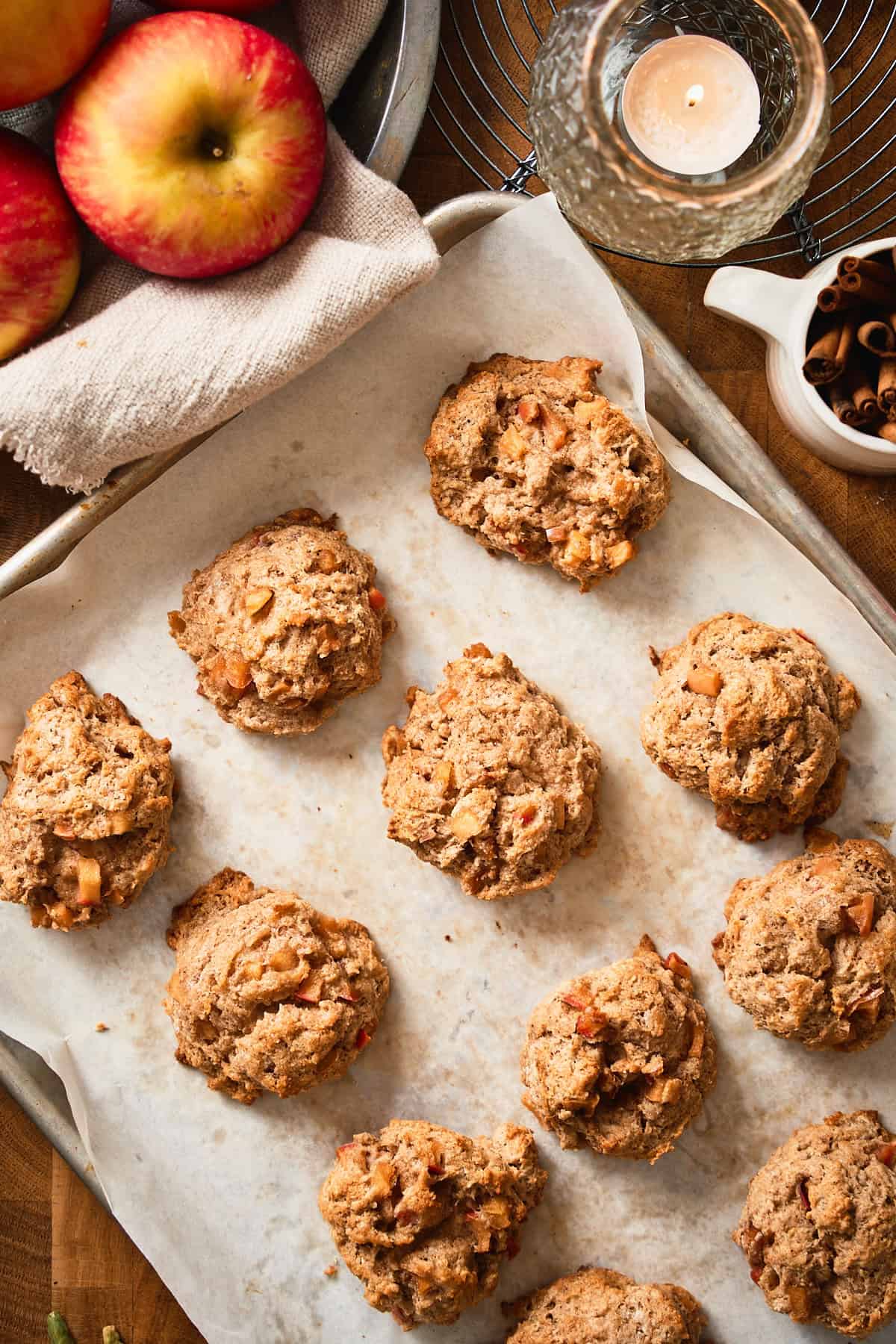 Baked apple biscuits on a baking sheet with cinnamon sticks, apples and twinkling tealight in the background.