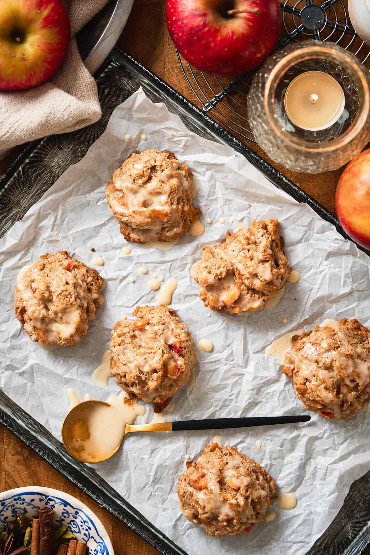 Baked apple drop biscuits on a parchment lined baking sheet drizzled with glaze with apples and candles surrounding.