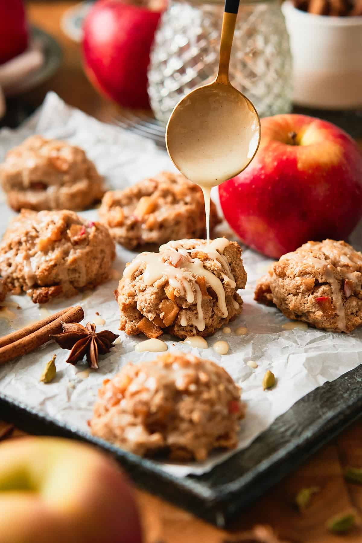 Beautiful shot of biscuits on a baking sheet with white parchment paper, surrounded by whole spices, fresh apples, and a drizzle of glaze going onto the biscuits.