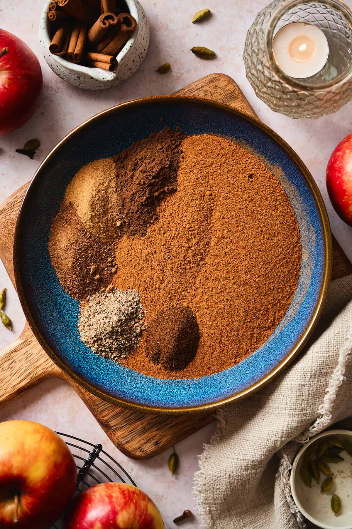 Ground spices measured into a blue bowl before mixing, surrounded by apples, cinnamon sticks, and cardamom pods.
