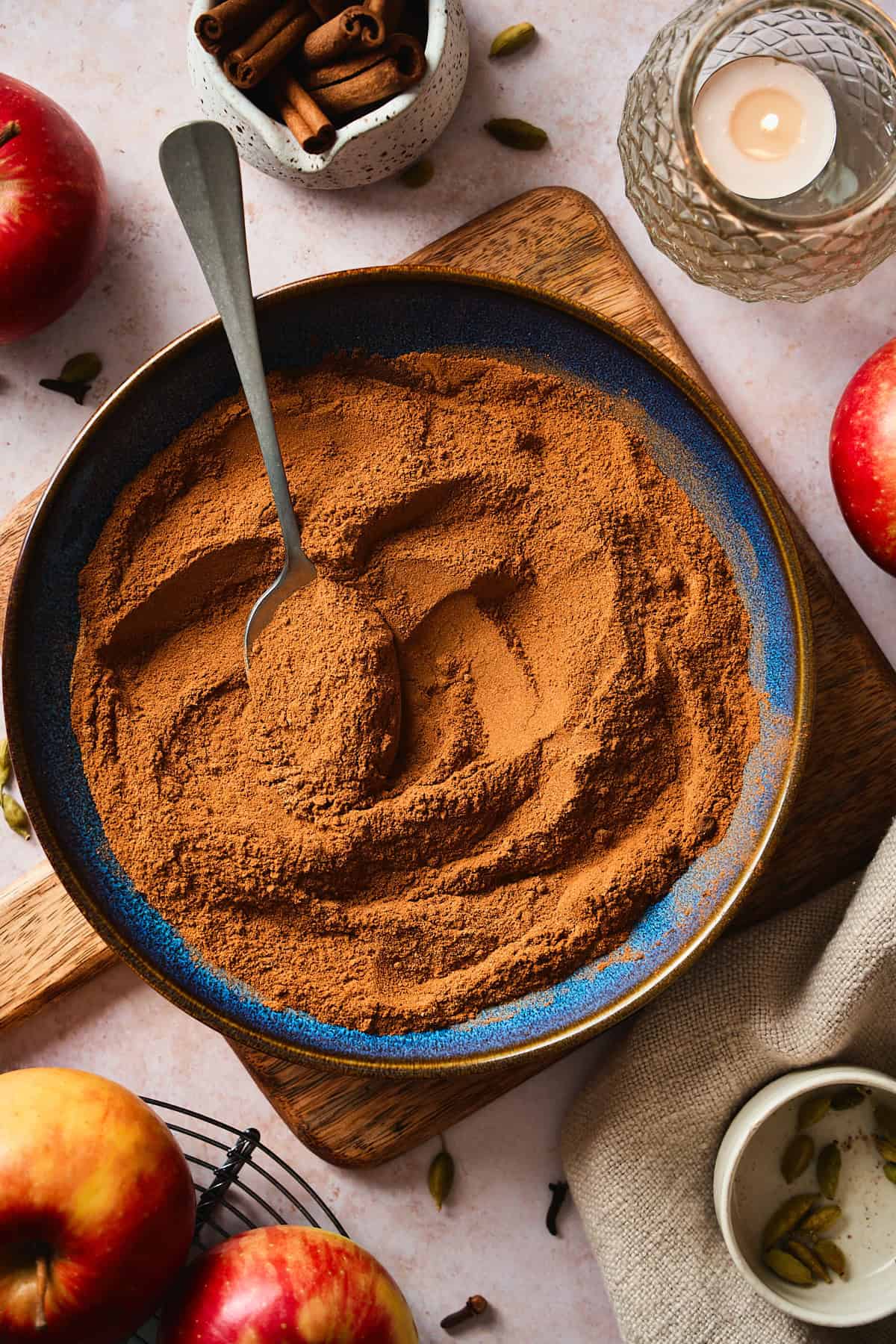Apple pie spice in a blue bowl with a spoon, styled with apples, cinnamon sticks, and a candle.