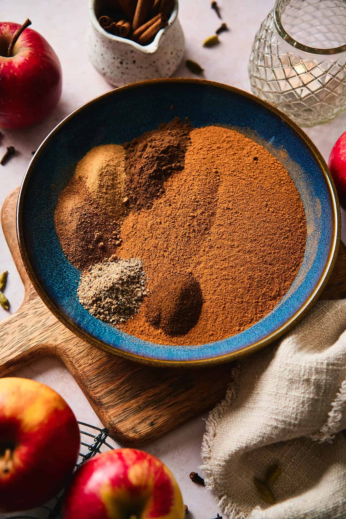 Angled view of ground spices measured into a blue bowl before mixing, with apples and cinnamon sticks in the background.
