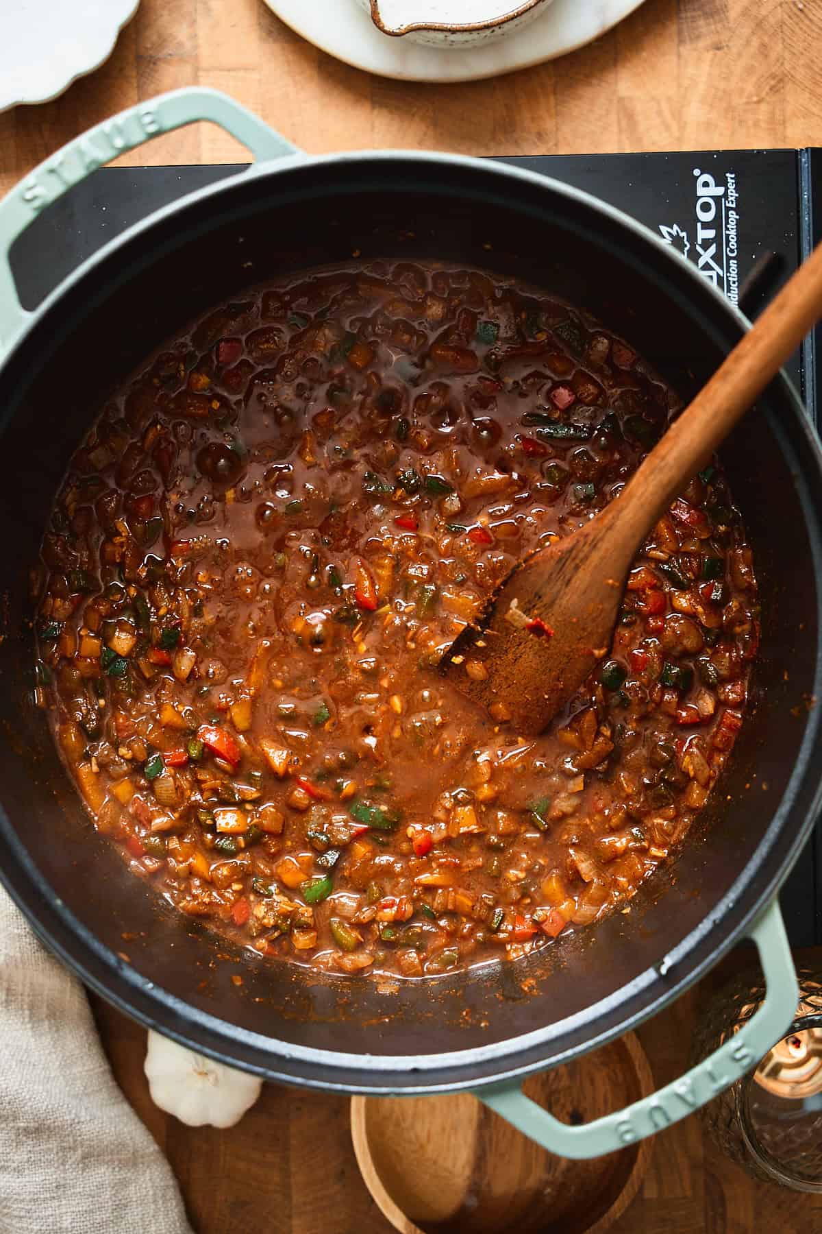 Pot with bell peppers and aromatics in a deep broth being stirred with a wooden spoon.