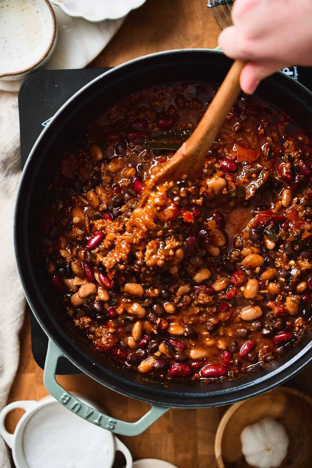 Hand stirring thick beer chili with a wooden spoon, showing beans, beef, and peppers.
