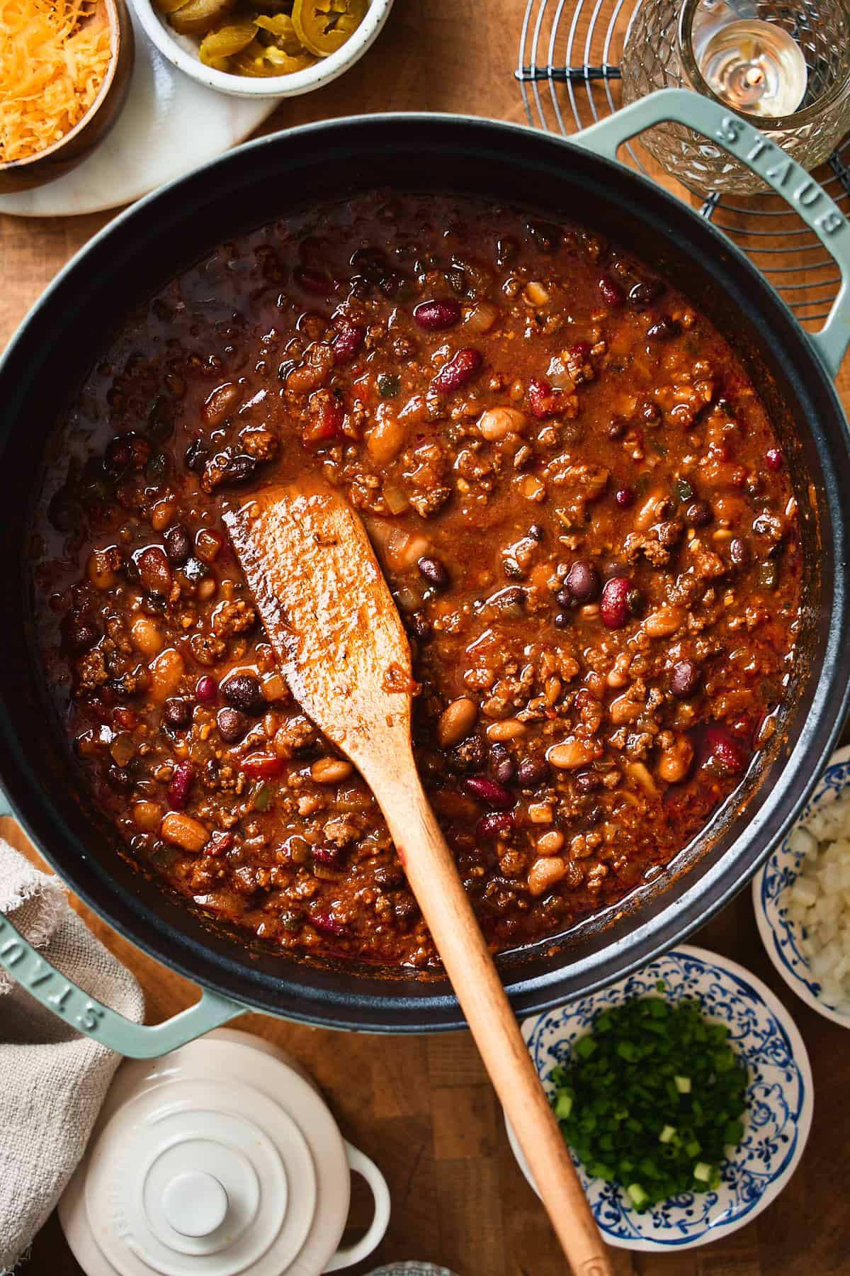 Finished beer chili in a Dutch oven surrounded by cheese, jalapeños, and green onions.