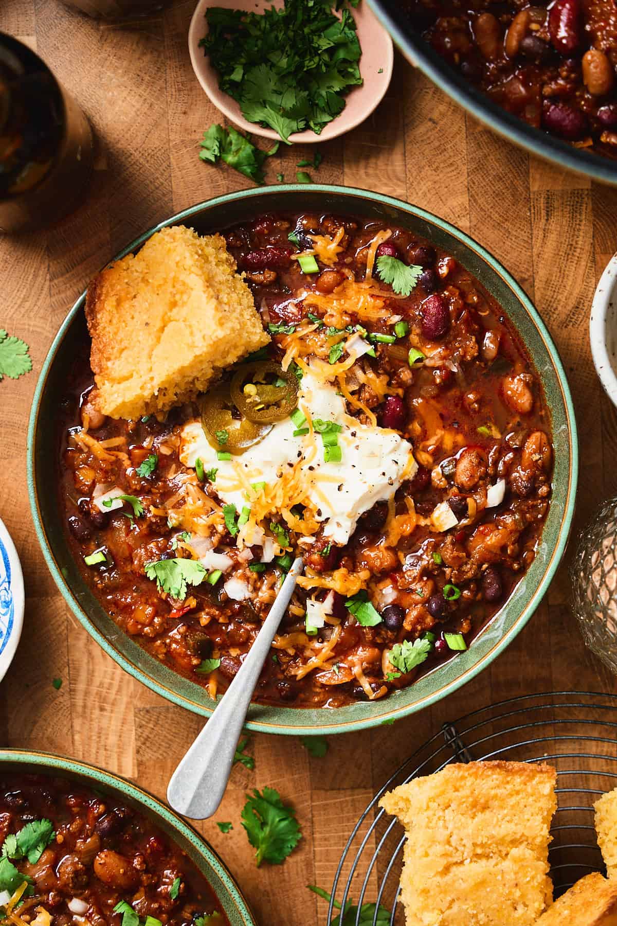 Bowl of dark beer chili topped with sour cream, cheese, jalapeños, and cornbread with a glass of dark beer nearby.