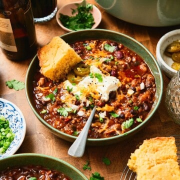 Close up shot of a green bowl of chili with beer, cornbread and toppings nearby.