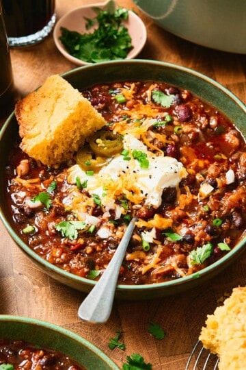 Close up shot of a green bowl of chili with beer, cornbread and toppings nearby.