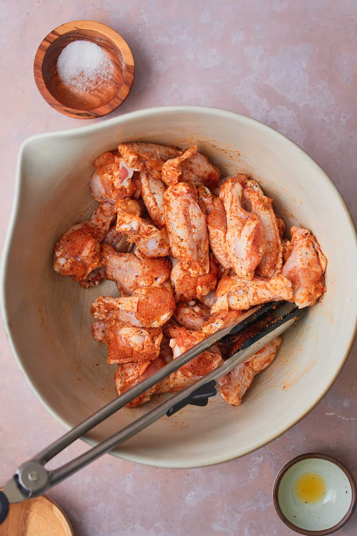 Raw chicken wings in a bowl tossed with seasonings, with tongs in the bowl.