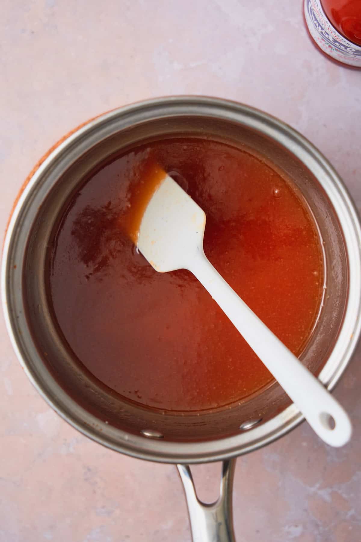 Red garlic butter honey sauce in a saucepan with a small rubber spatula in the pan.