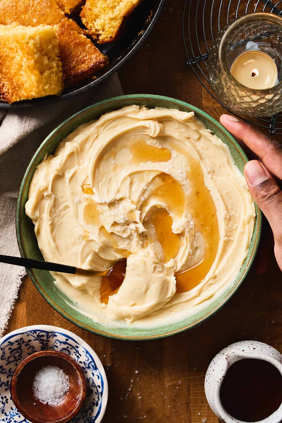 Hand reaching for a bowl of fluffy whipped maple butter drizzled with maple syrup, styled with cornbread and a candle.