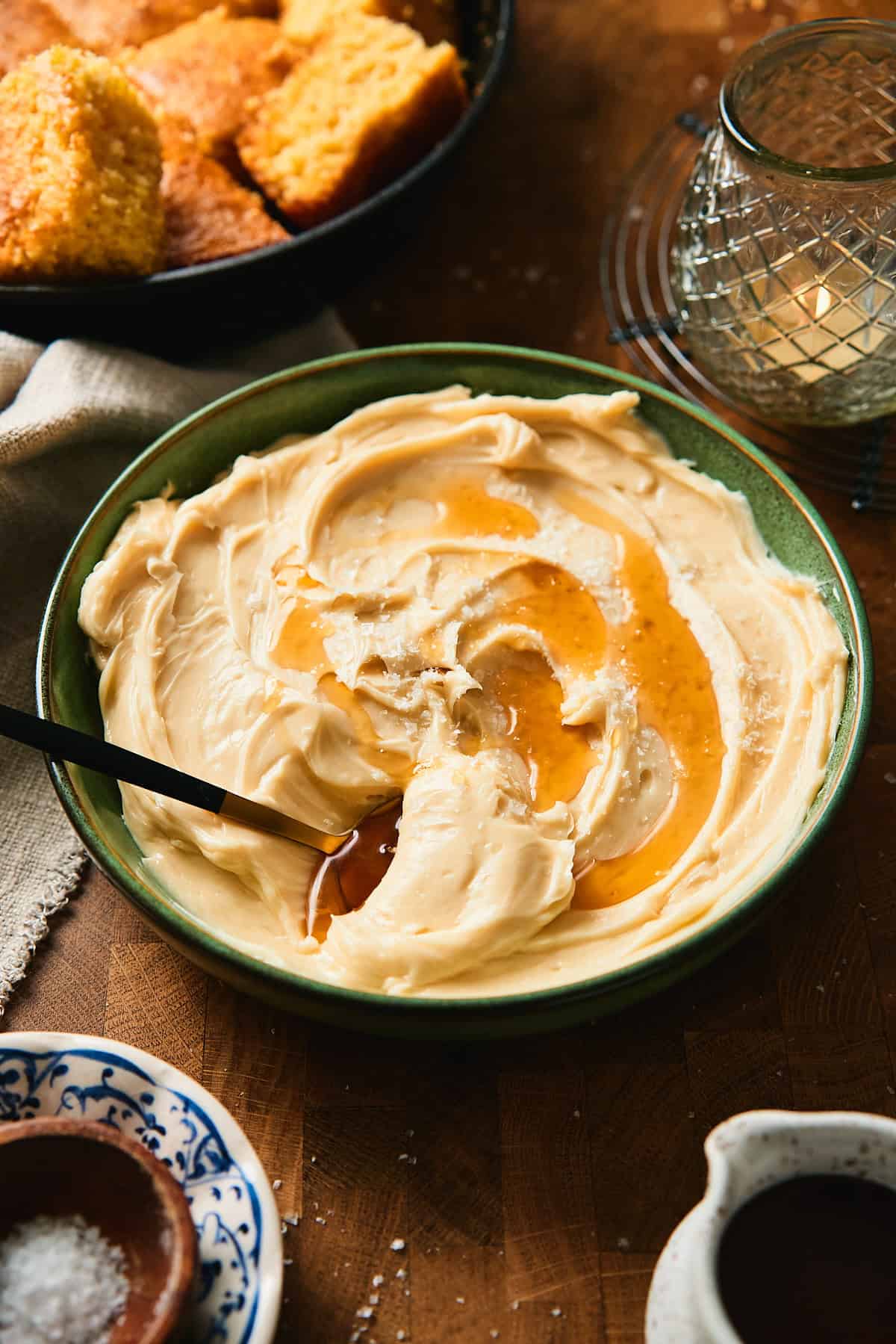 Close-up of whipped maple butter swirled in a green bowl with maple syrup and a knife, cornbread in the background.