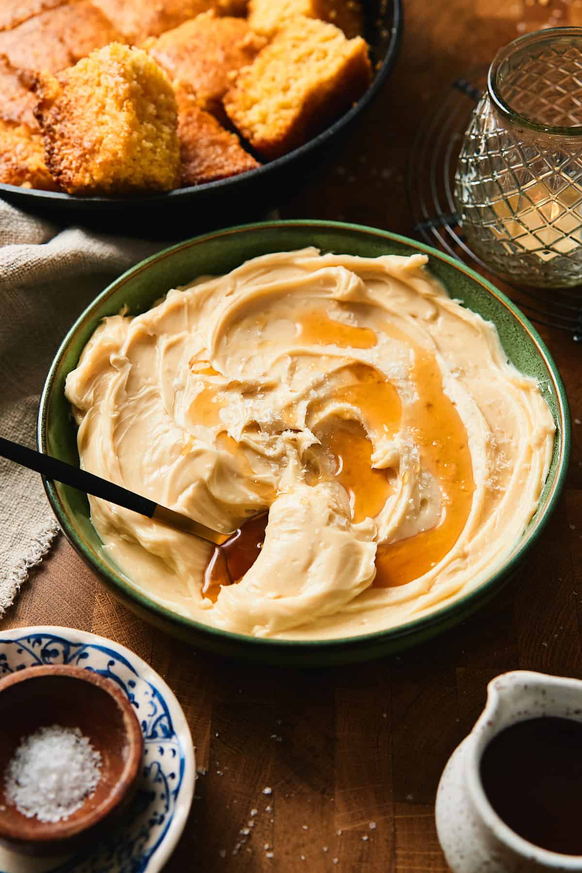 Bowl of whipped maple butter with pooled maple syrup, knife, cornbread, salt, and candle on a wooden table.