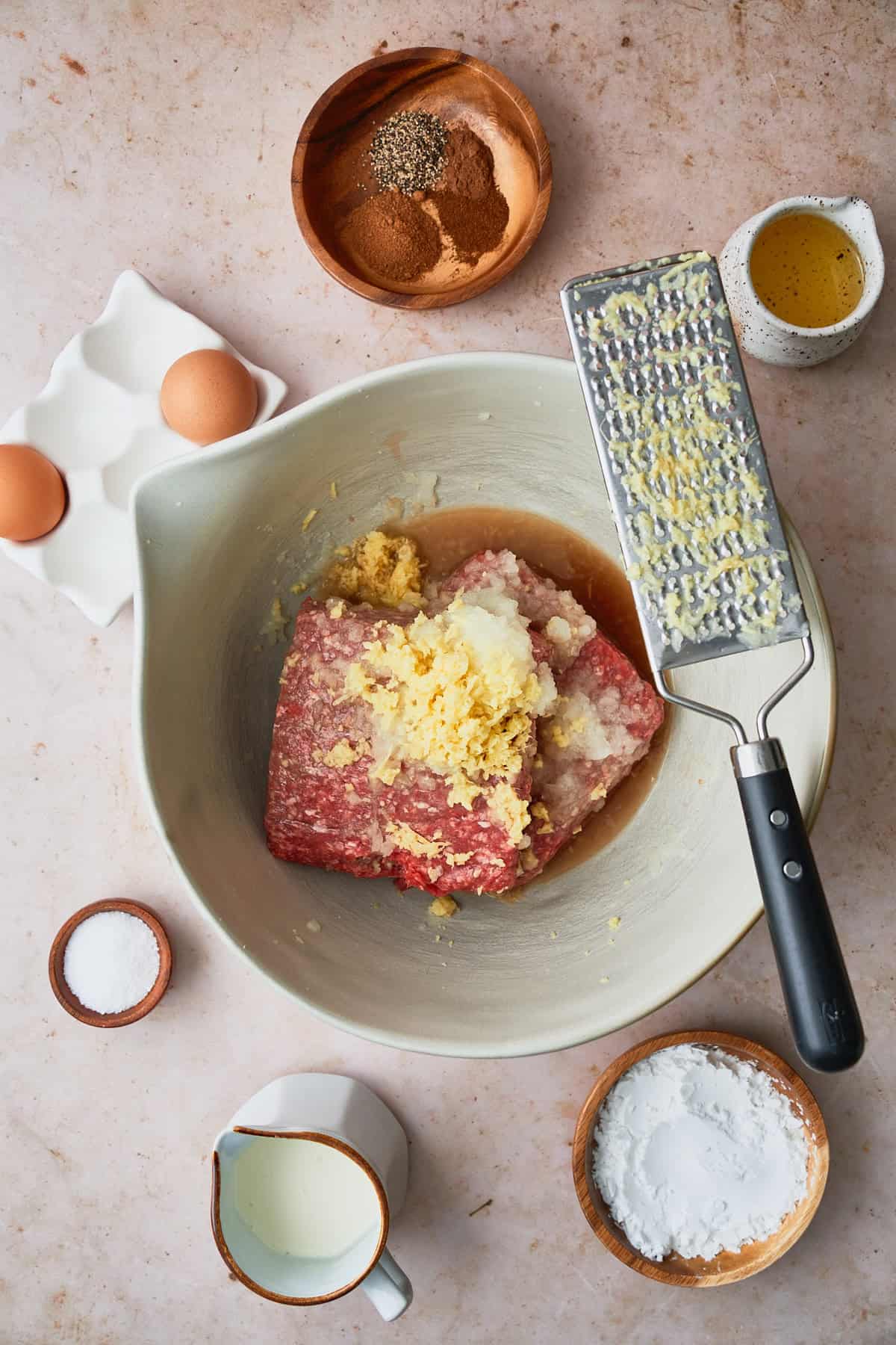 Bowl of raw ground beef with a grater over top, with onion and ginger grated into the bowl, surrounded by eggs, salt, starch, spices, oil, and other ingredients to make the meatballs.