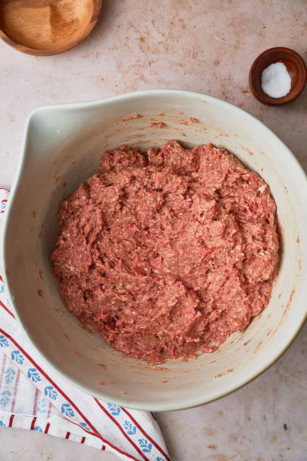 Raw ground beef mixture to make meatballs in a large bowl with a cute Nordic inspired towel underneath, with salt nearby.