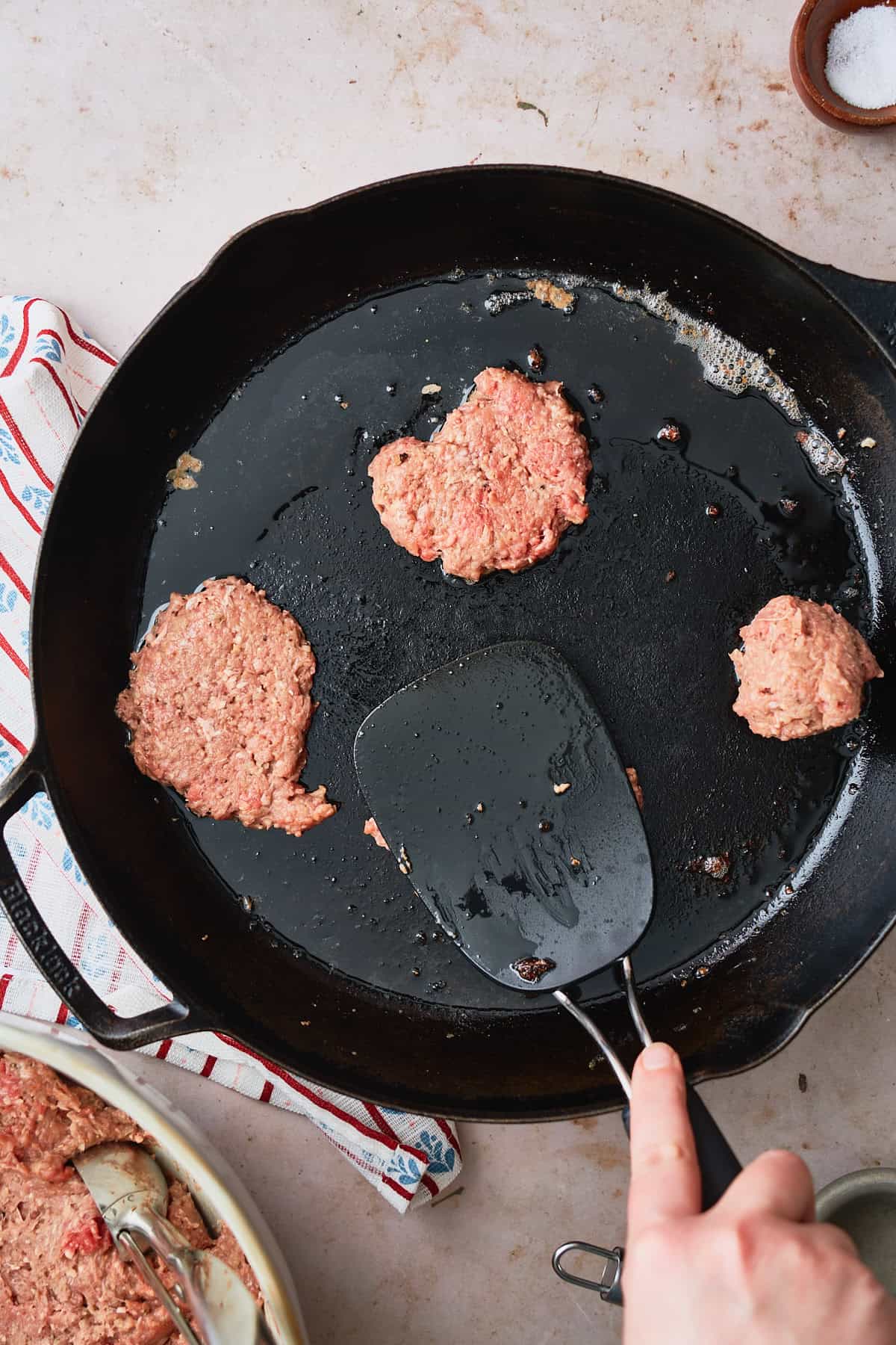 Hand pressing meat patties flat in a hot cast iron skillet with a Nordic napkin underneath.