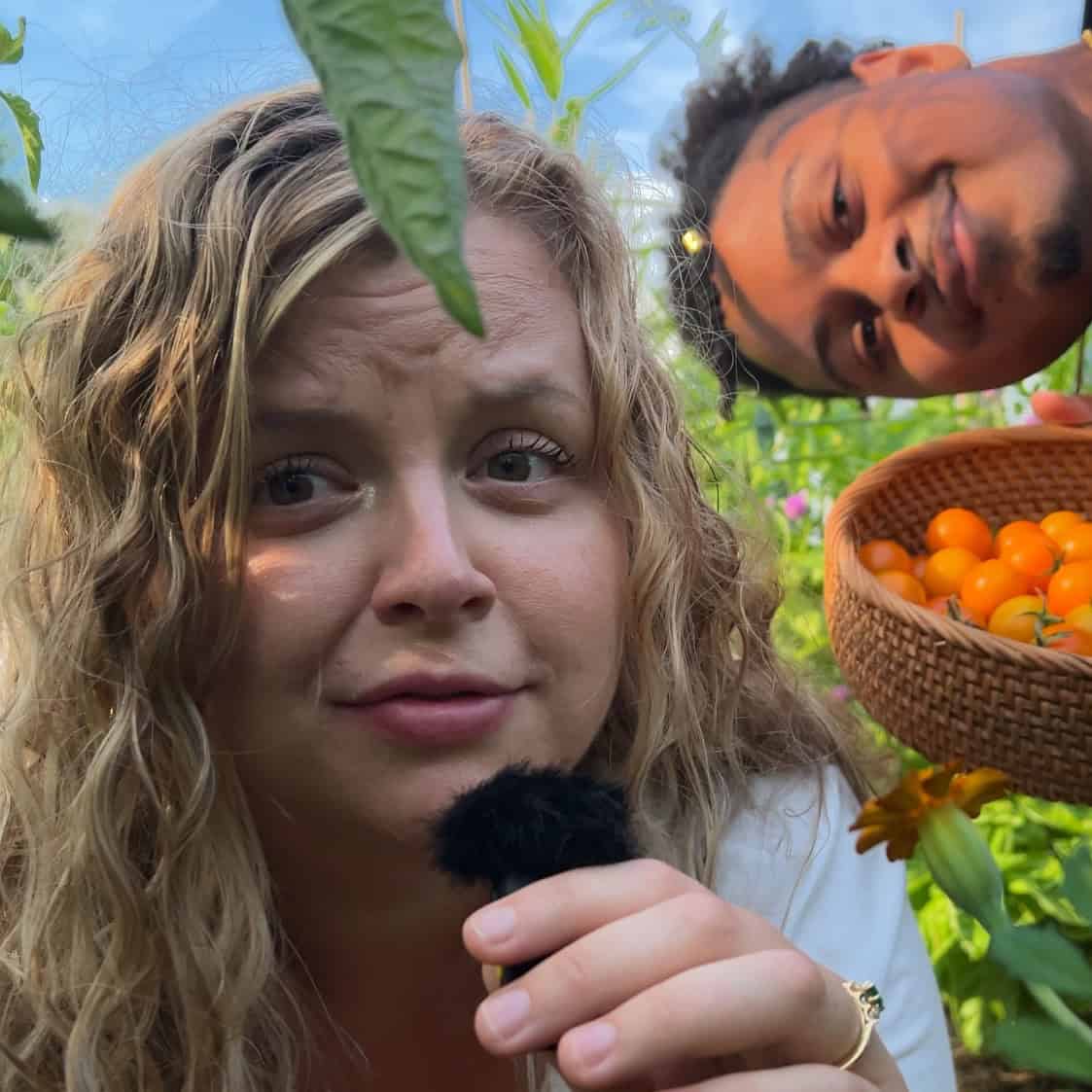 husband and wife of A Full Living in their garden with a basket of sungold tomatoes and a microphone.